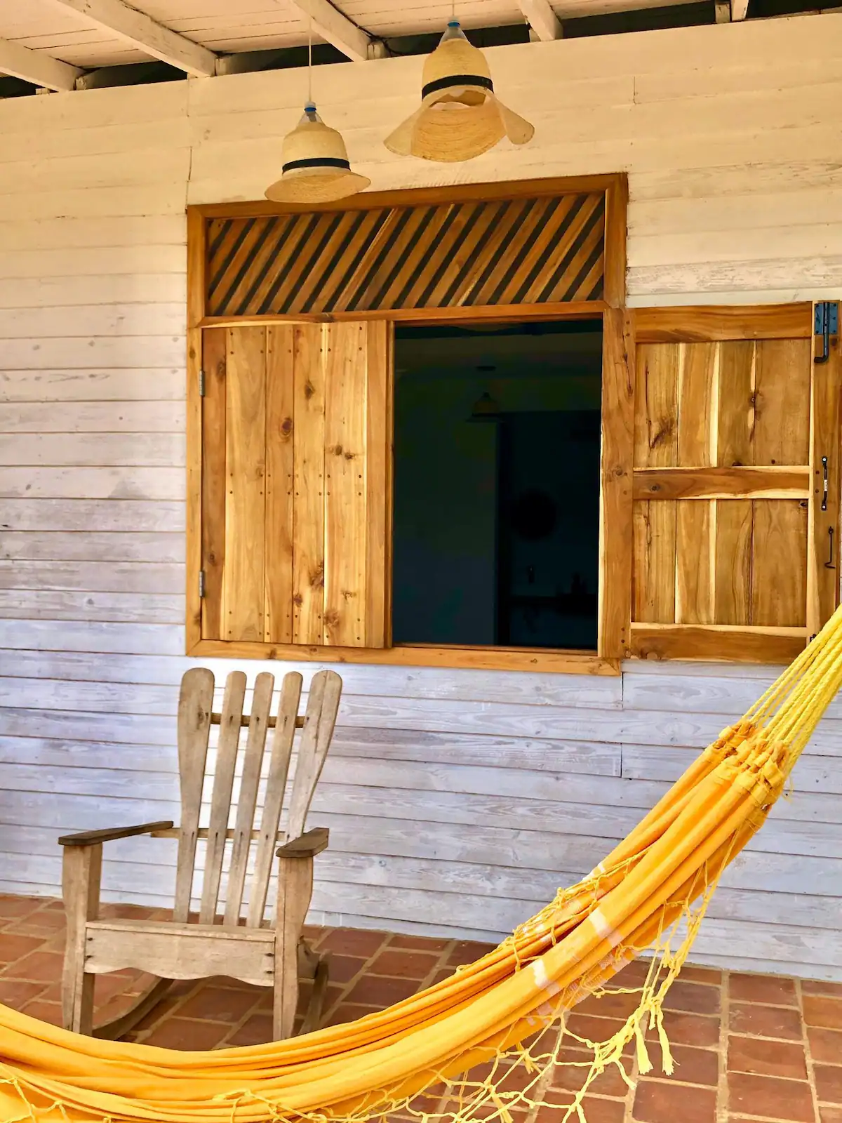A wooden porch scene featuring a hanging hammock chair, a wooden rocking chair, and a window with wooden shutters and a rolling vent above it.