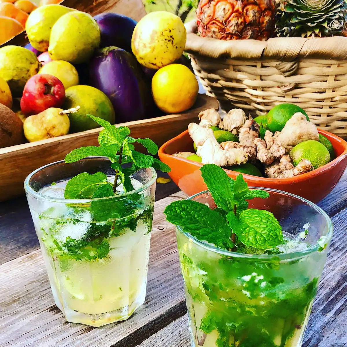 Two glasses of mojito with fresh mint leaves on a wooden table, surrounded by a basket of pineapple, ginger, lime, and other fresh fruits.