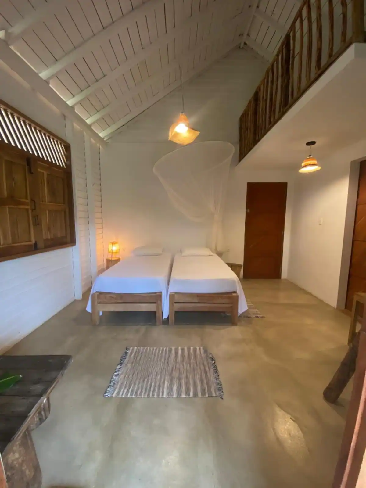 A bedroom with two beds pushed together, a mosquito net hanging above, and wooden accents, in a rustic style with a high vaulted ceiling.