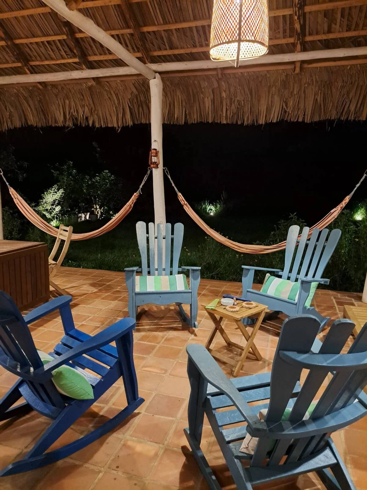 Nighttime outdoor patio with four blue wooden chairs, a small wooden table with snacks, and a hammock under a thatched roof, surrounded by greenery.