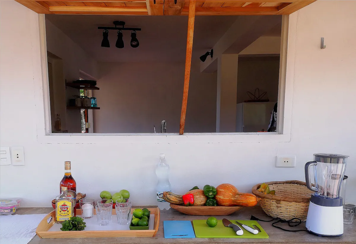 Kitchen counter with a tray of limes and a bottle of alcohol, a bowl of vegetables, a blender, and kitchen tools in front of a window opening to the kitchen interior.