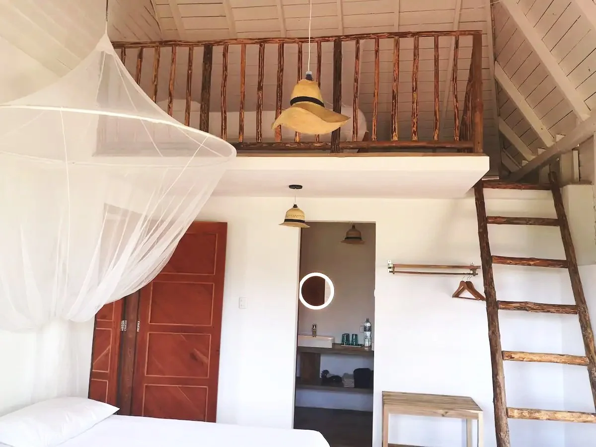 A cozy bedroom with white walls, a bed with a canopy, a wooden wardrobe, and a small loft area accessible by a ladder, featuring rustic wooden railing and a hat hanging from a light fixture.