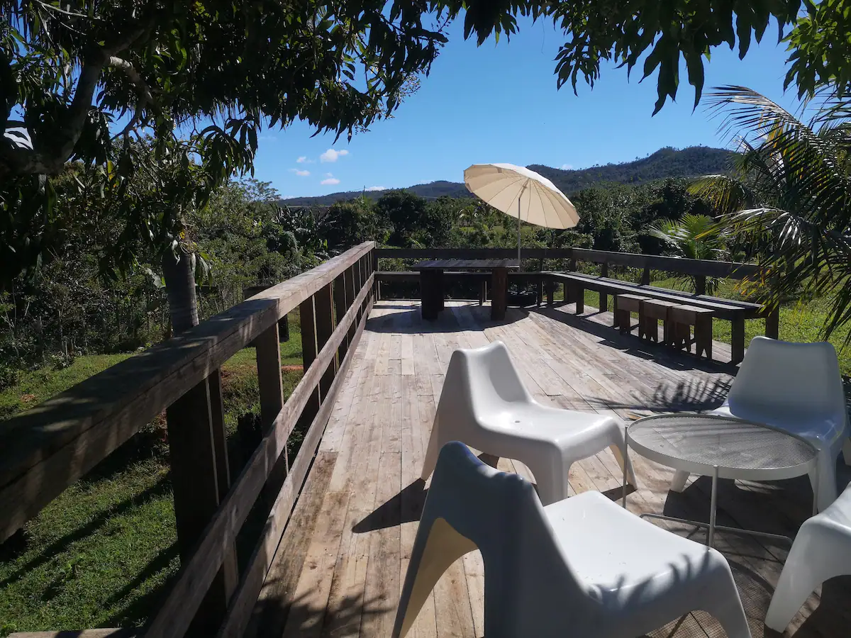 Deck with white chairs, glass-top table, beige umbrella, surrounded by lush green trees and mountains in the background under a blue sky.