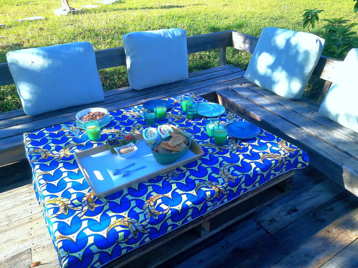 Outdoor dining setup with a blue and white patterned tablecloth, on a wooden table with cushions, set for a meal with bowls of cereal, bread, cups of juice, and condiments.