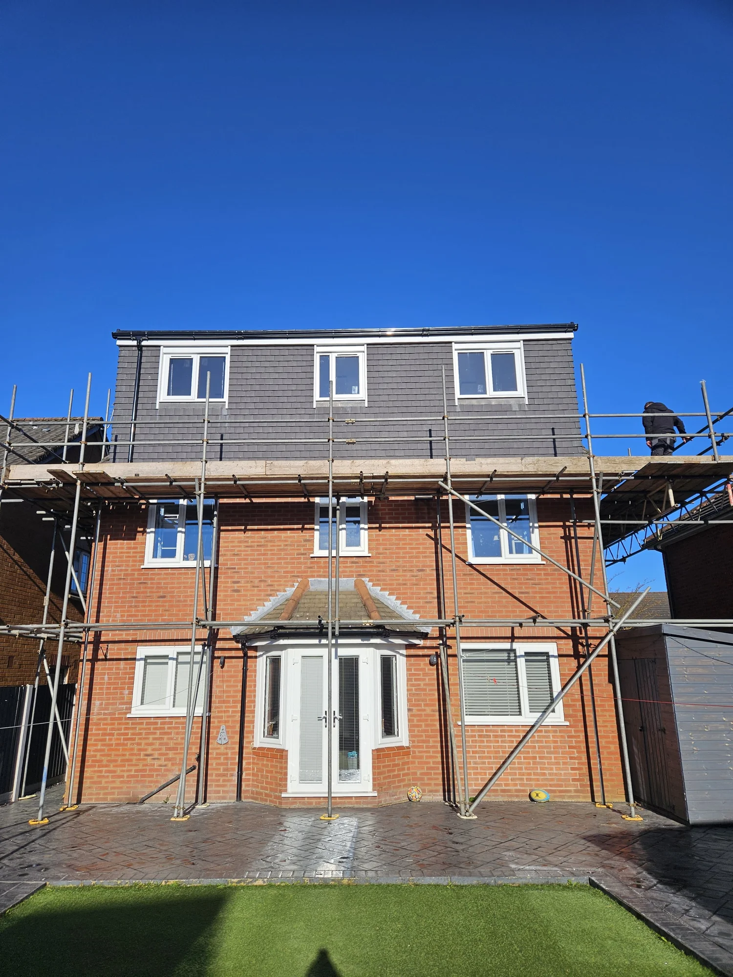 House under renovation with scaffolding, brick base and black roof against a clear blue sky