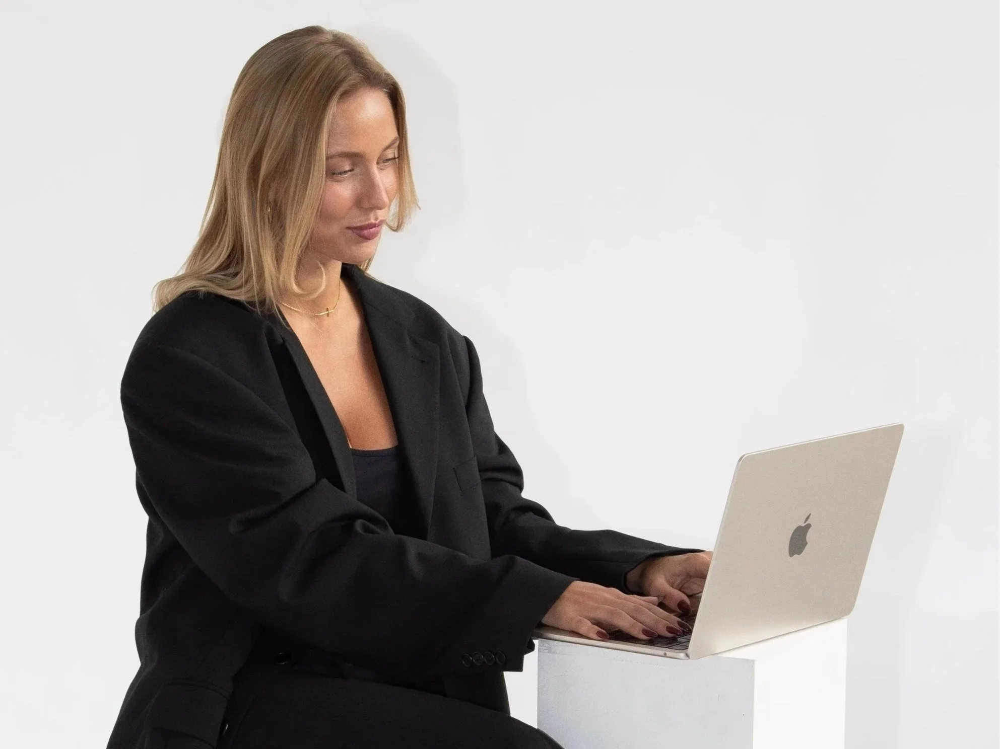 Kvinde i sort blazer arbejder på en MacBook mod en hvid baggrund.