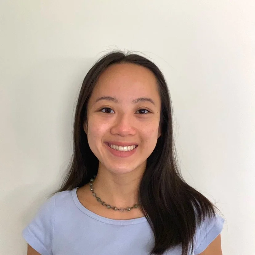A young woman with long dark hair, smiling, wearing a light purple T-shirt and a beaded necklace, standing against a plain white wall.