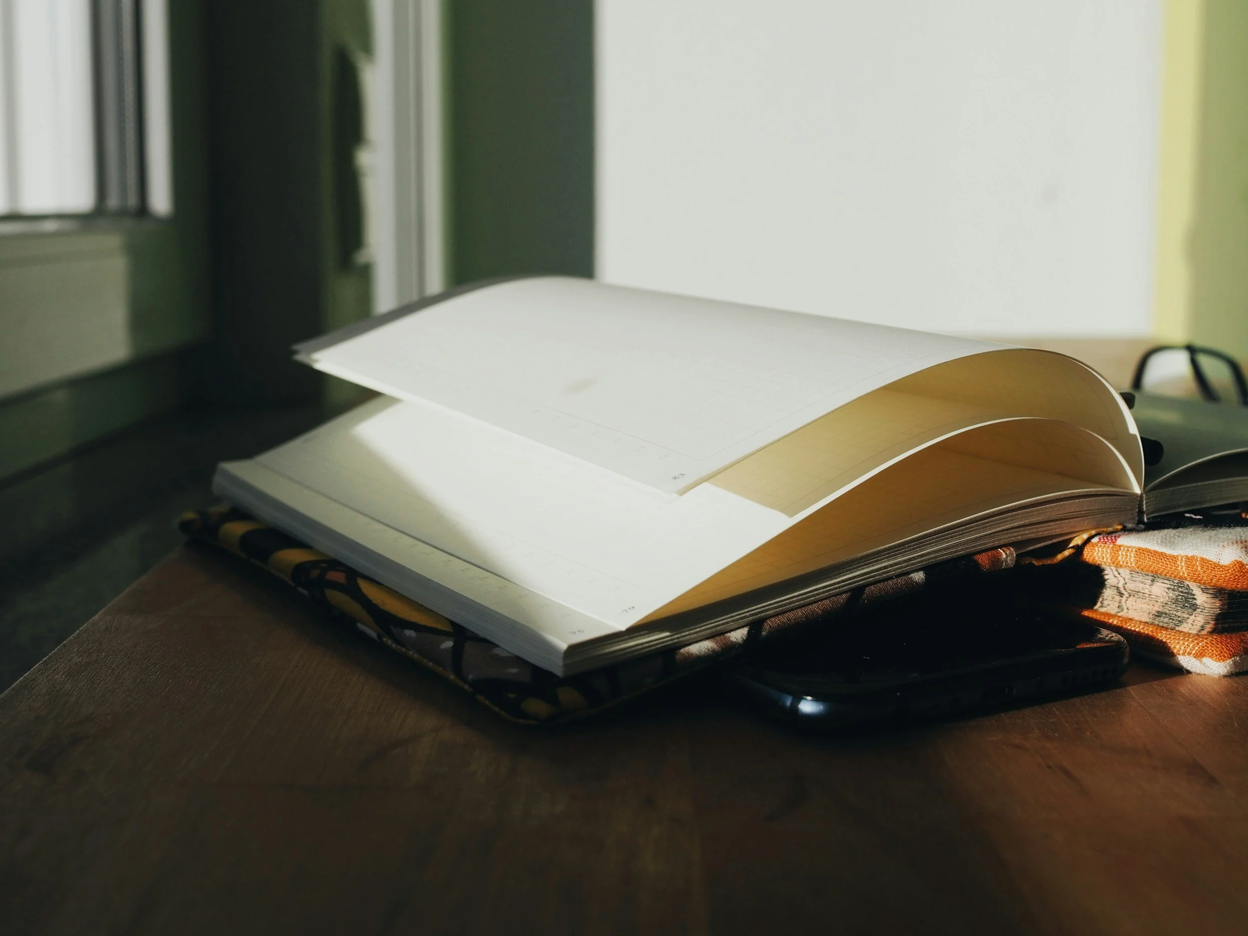 Un cahier ouvert avec des pages jaunes, posé sur une table en bois, avec un téléphone portable en dessous, près d'une fenêtre avec la lumière du soleil