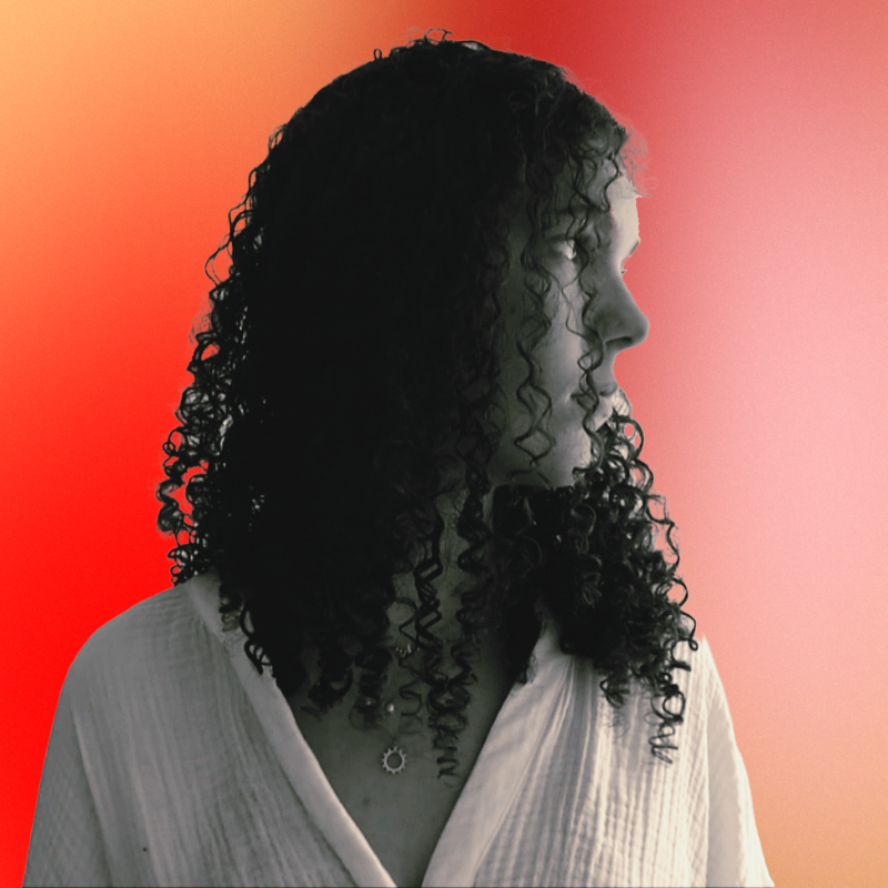 A woman with curly dark hair looking to the right, wearing a white shirt, with a blurred red to pink gradient background.