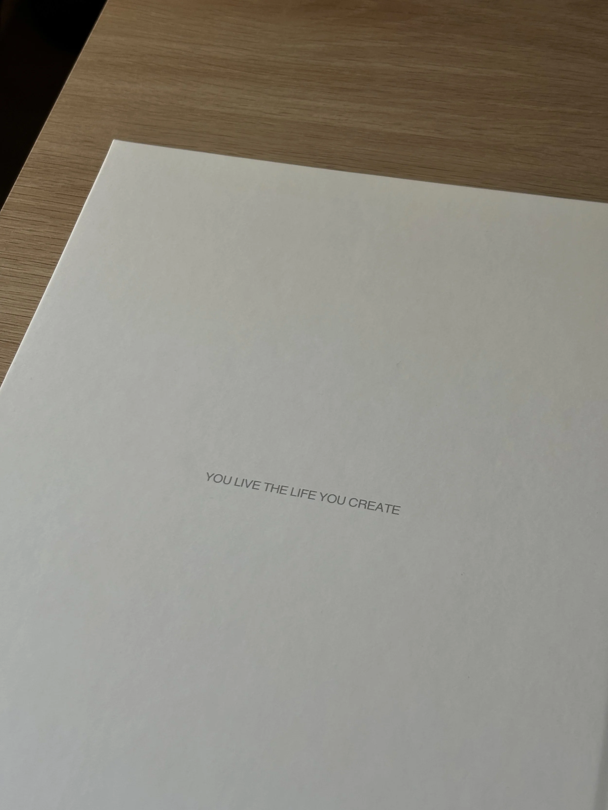Close-up of a paper with the phrase "YOU LIVE THE LIFE YOU CREATE" printed on it, placed on a light wooden surface.
