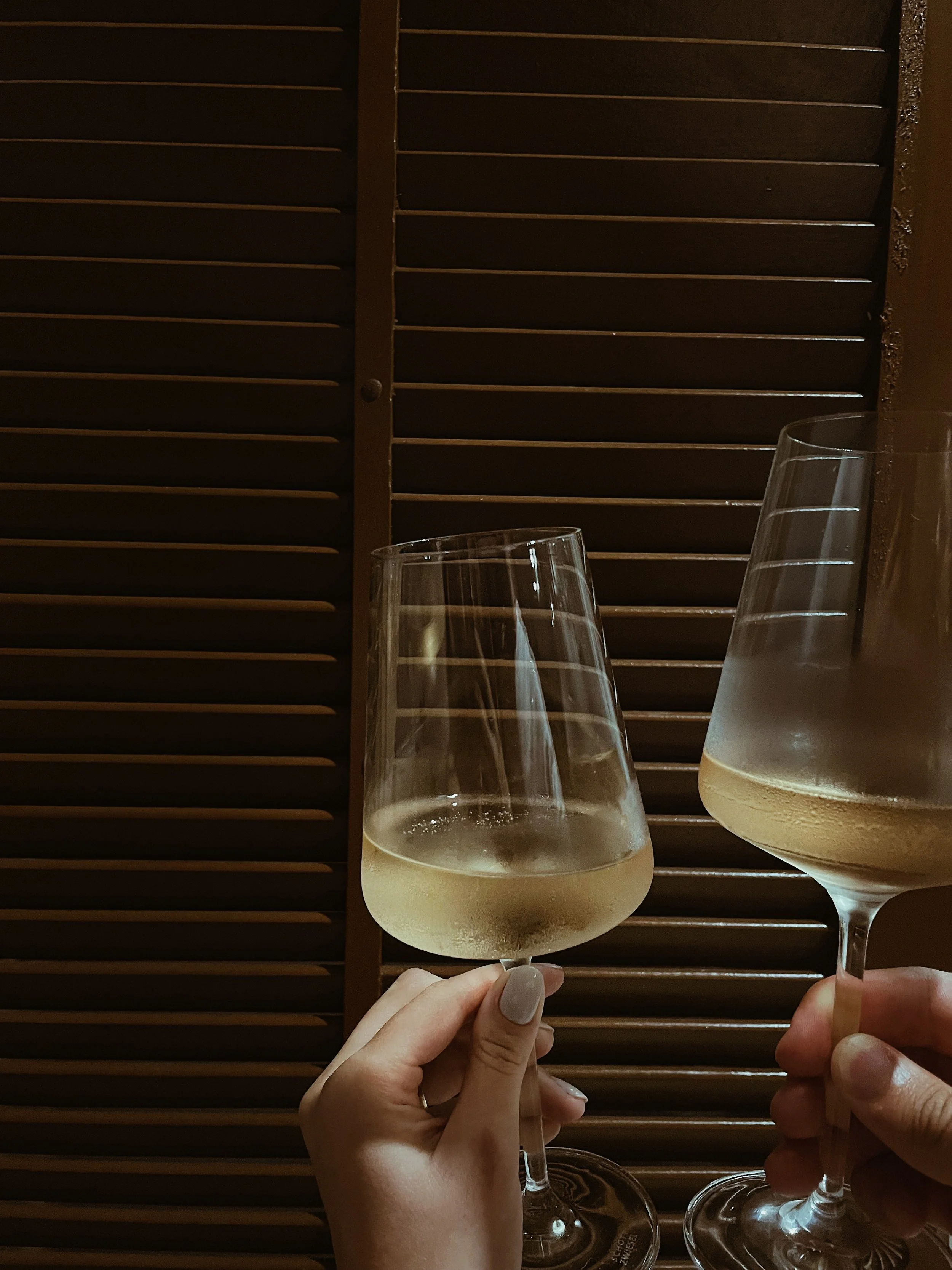 Two hands holding wine glasses with white wine, against a background of wooden blinds.