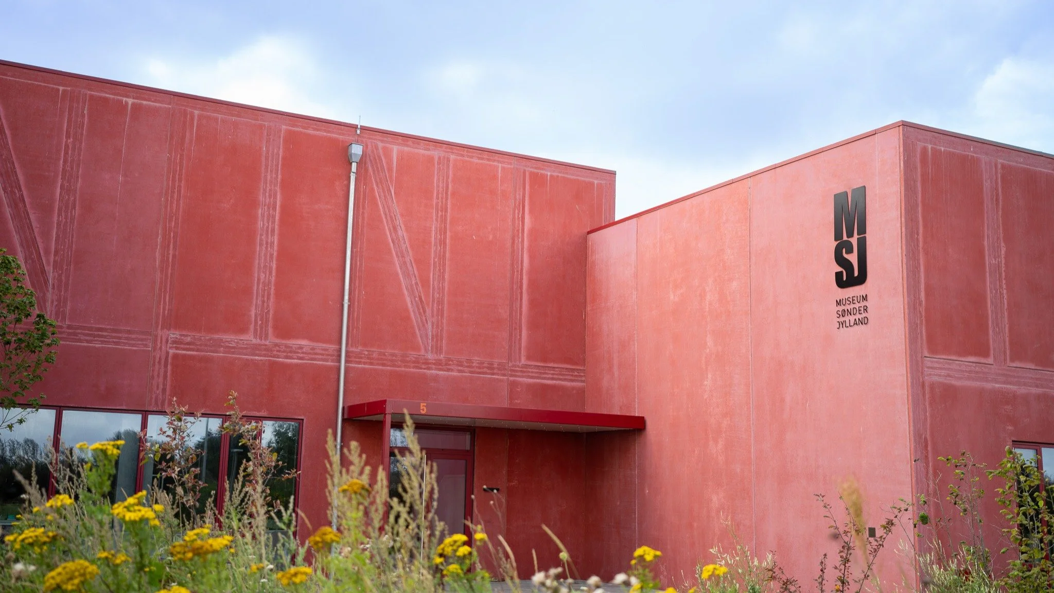 Exterior of Museum Sønderjylland building, featuring red walls and signage with MSJ initials, surrounded by wildflowers.