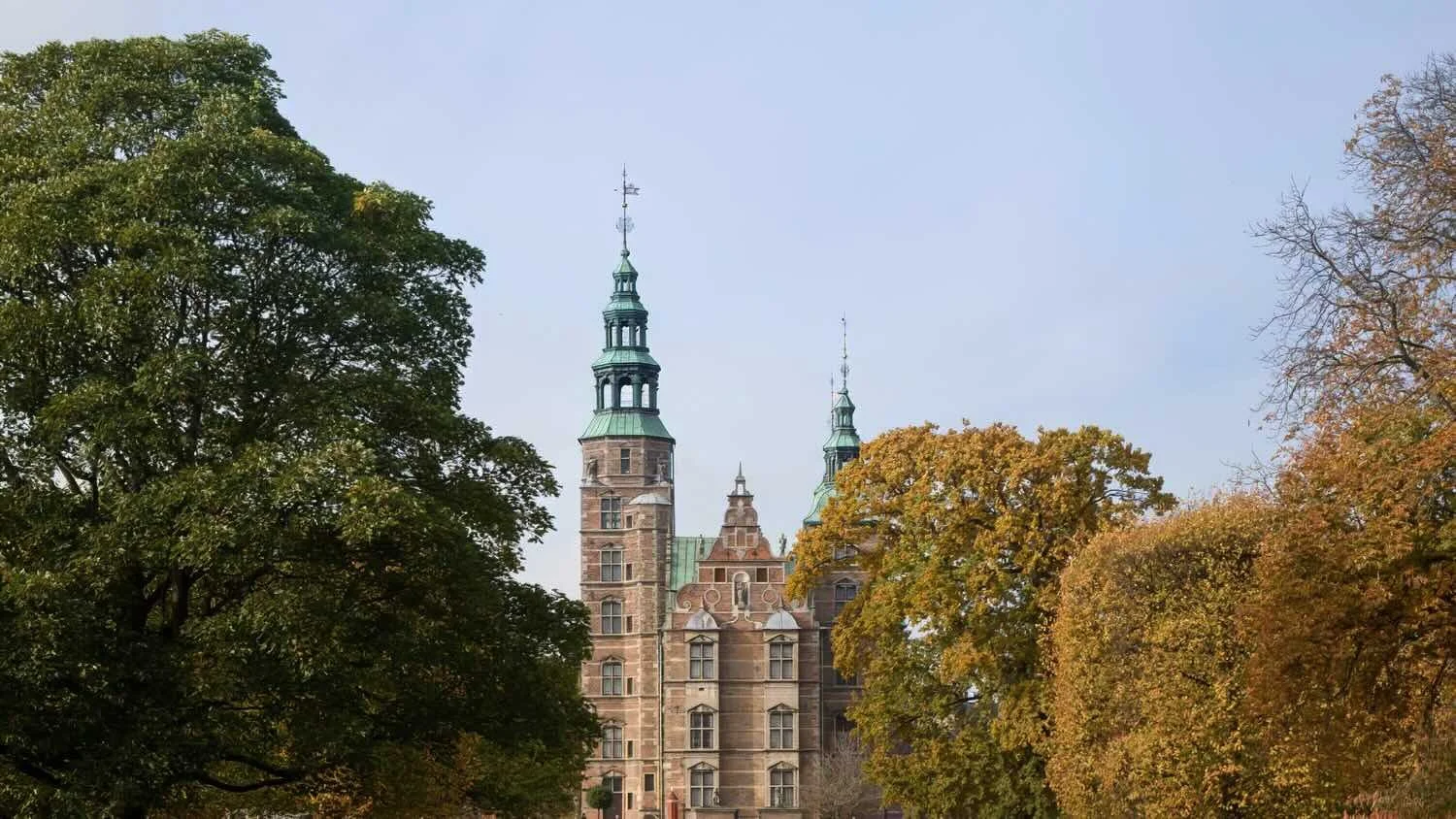 Historisk slotsbygning omgivet af træer med efterårsfarver mod en klar blå himmel.