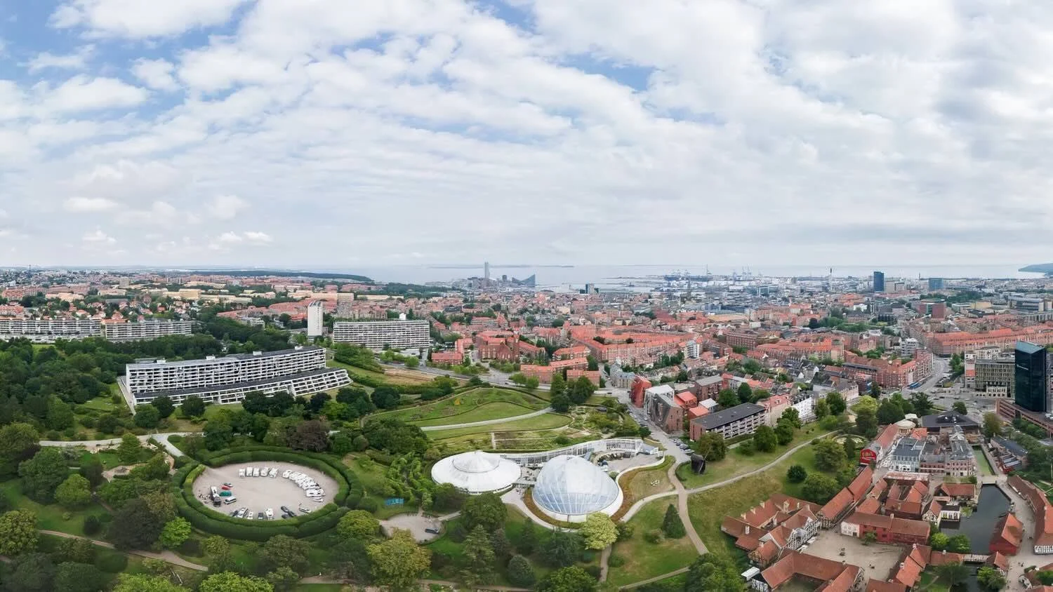 Wide aerial view of Aarhus, Denmark, showing the city, harbor, and the glass domes of the Aarhus Botanical Garden within a large park.