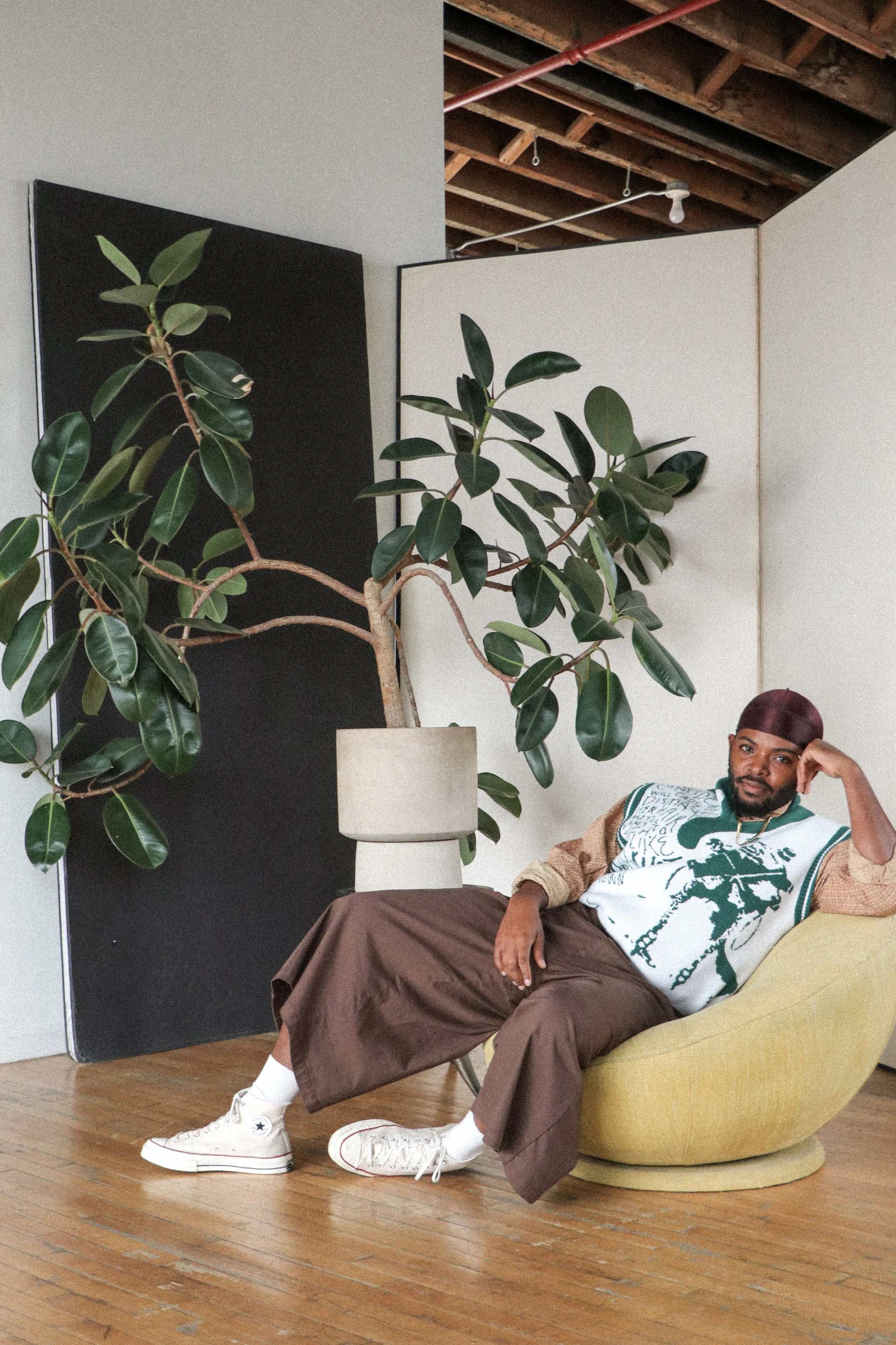 A photo of Keanu sitting on a beanbag chair in front of a large rubber plant. He is wearing a green and brown outfit and looks at the camera peacefully.