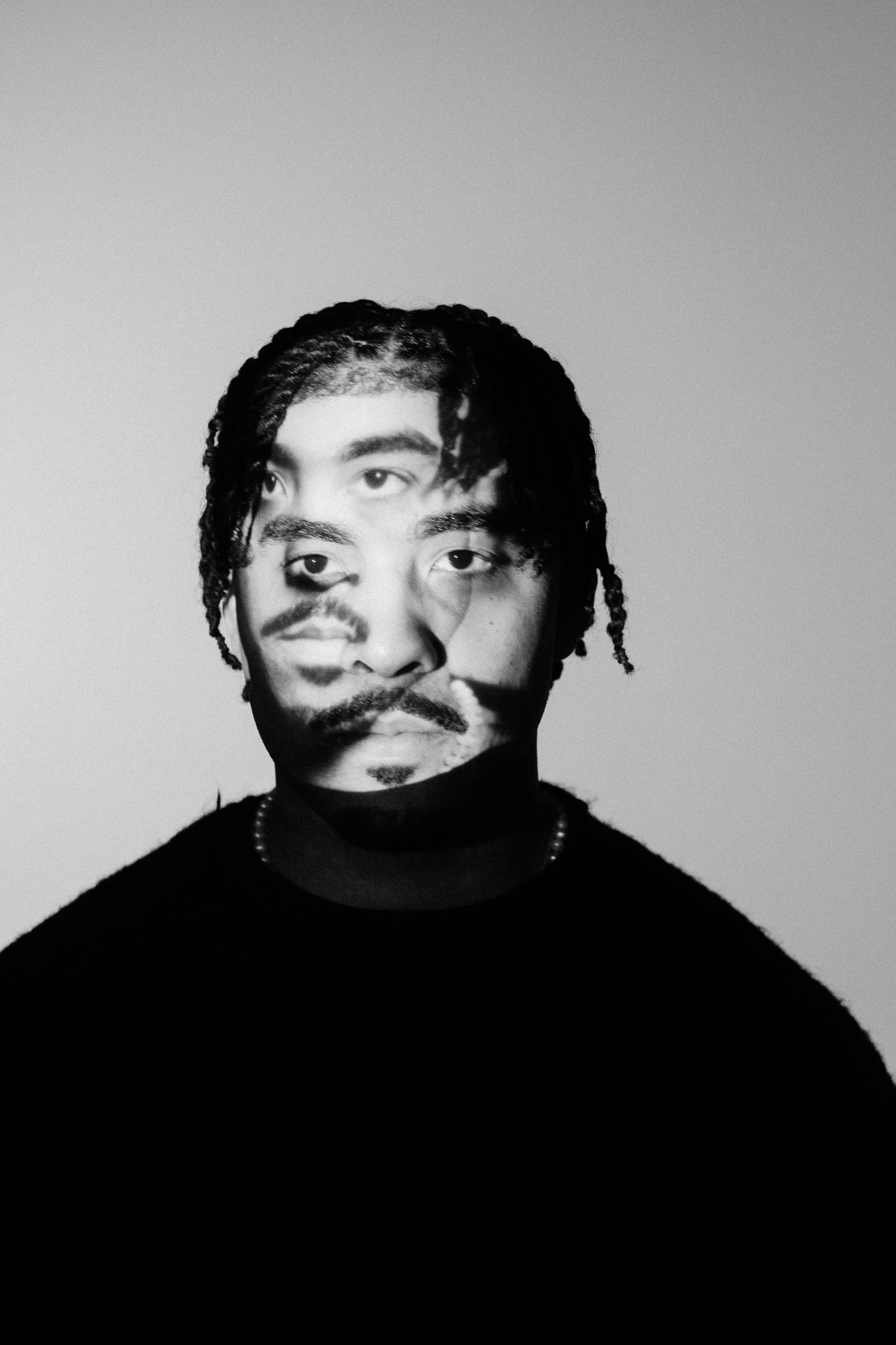 An artistic black and white image of a Black man in twists with multiple projections of his face on top of his face