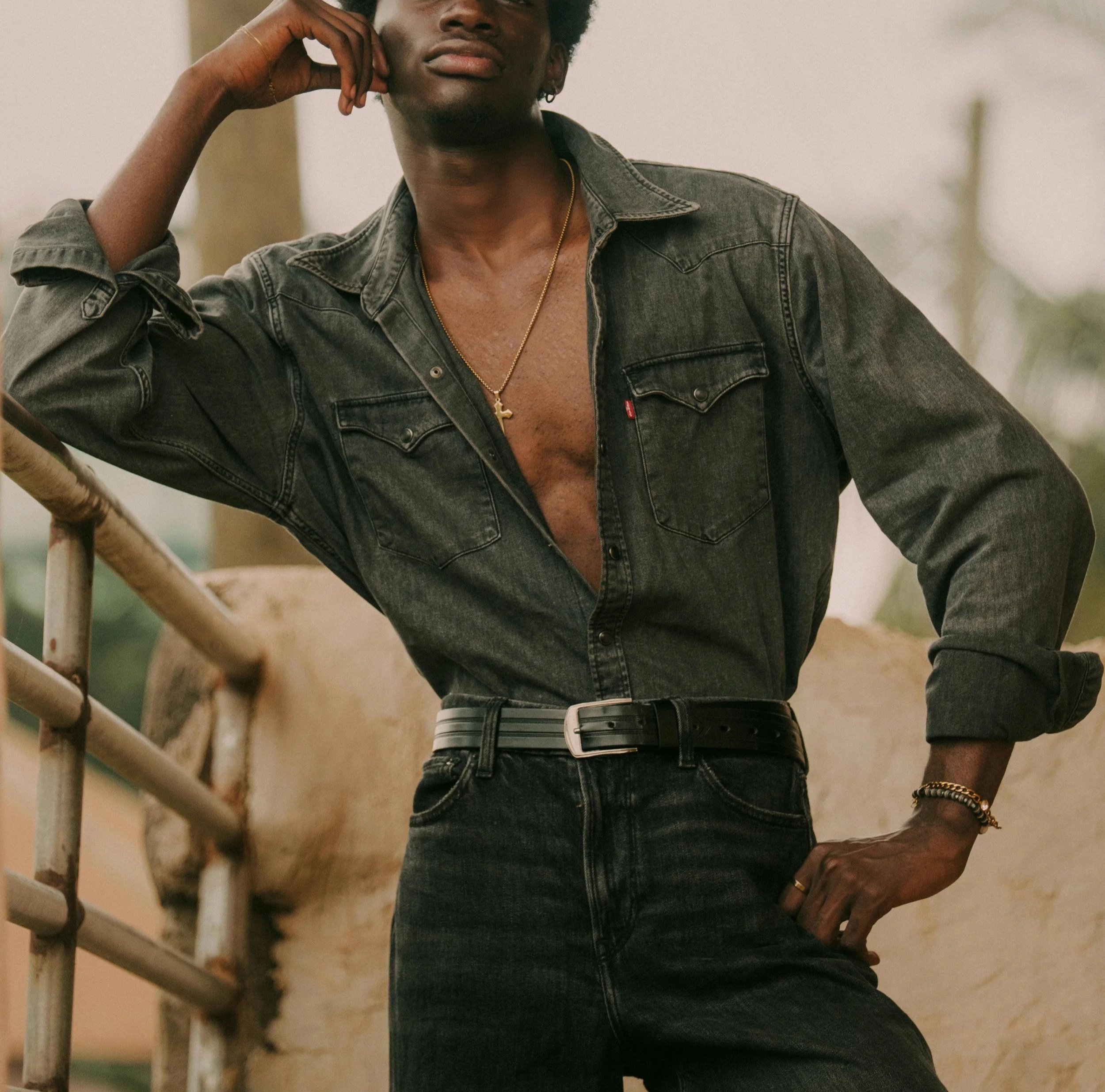 A Black man in an open denim shirt and belt poses in front of the camera
