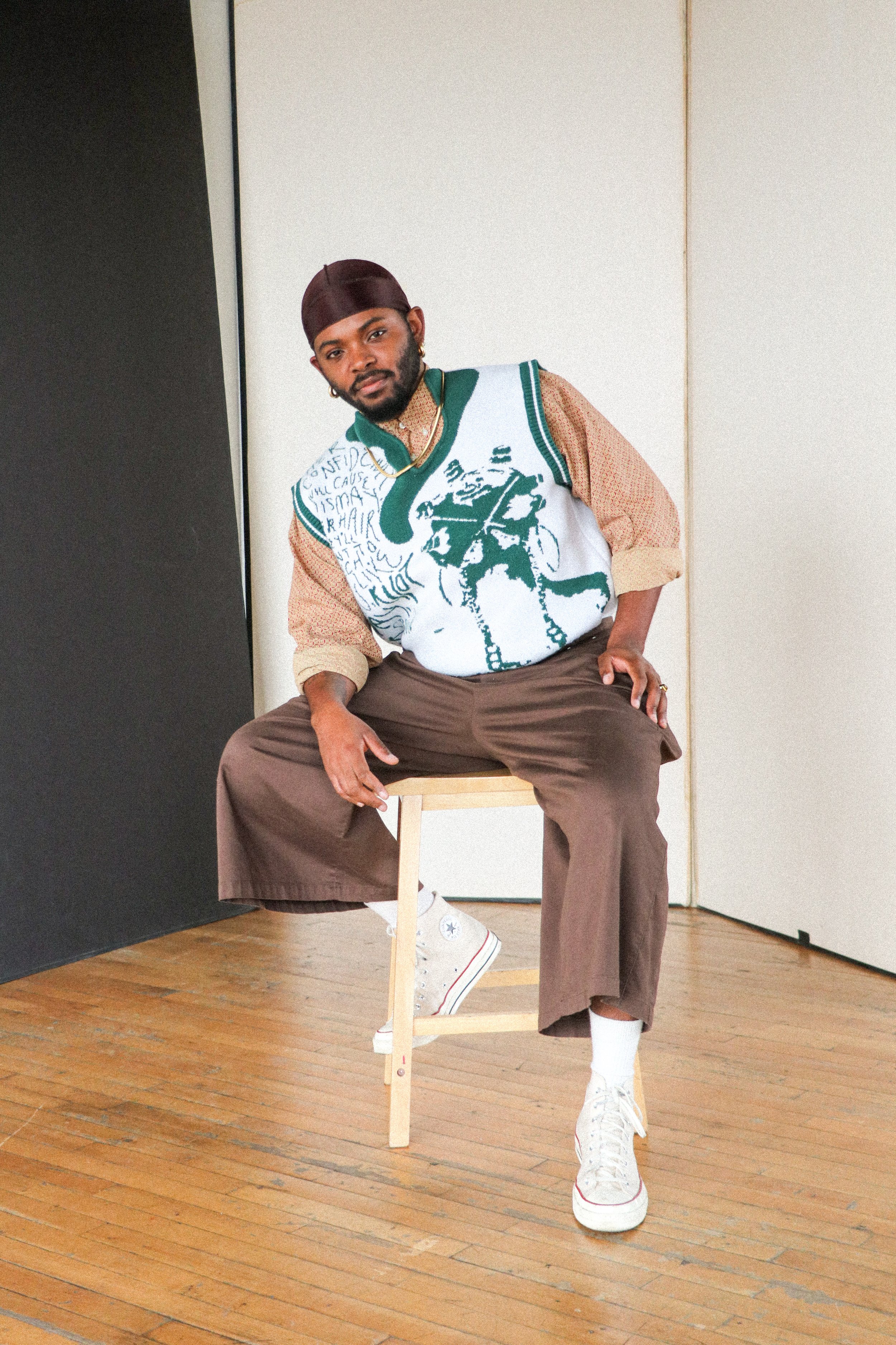 A man sitting on a wooden stool in a simple room with hardwood floors and plain walls, wearing a brown bandana, artwork-designed sweater vest, beige shirt, brown wide-leg pants, white high-top Converse sneakers, and accessorized with gold jewelry.