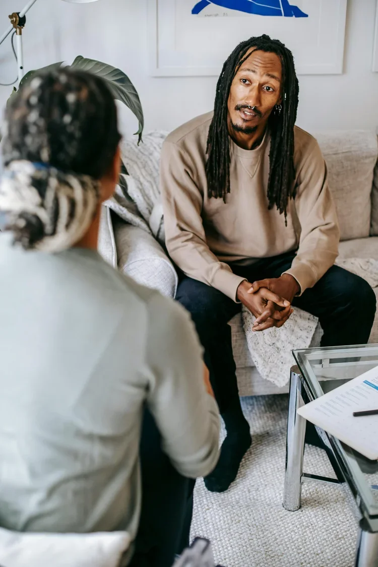 An image of a Black man sitting in front of a therapist in braids, his hands folded pensively