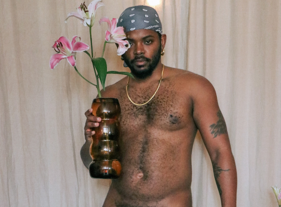 Keanu Jackson LCSW stands shirtless in front of a white curtain. He's wearing a gray bandana and holds a vase of pink lillies.