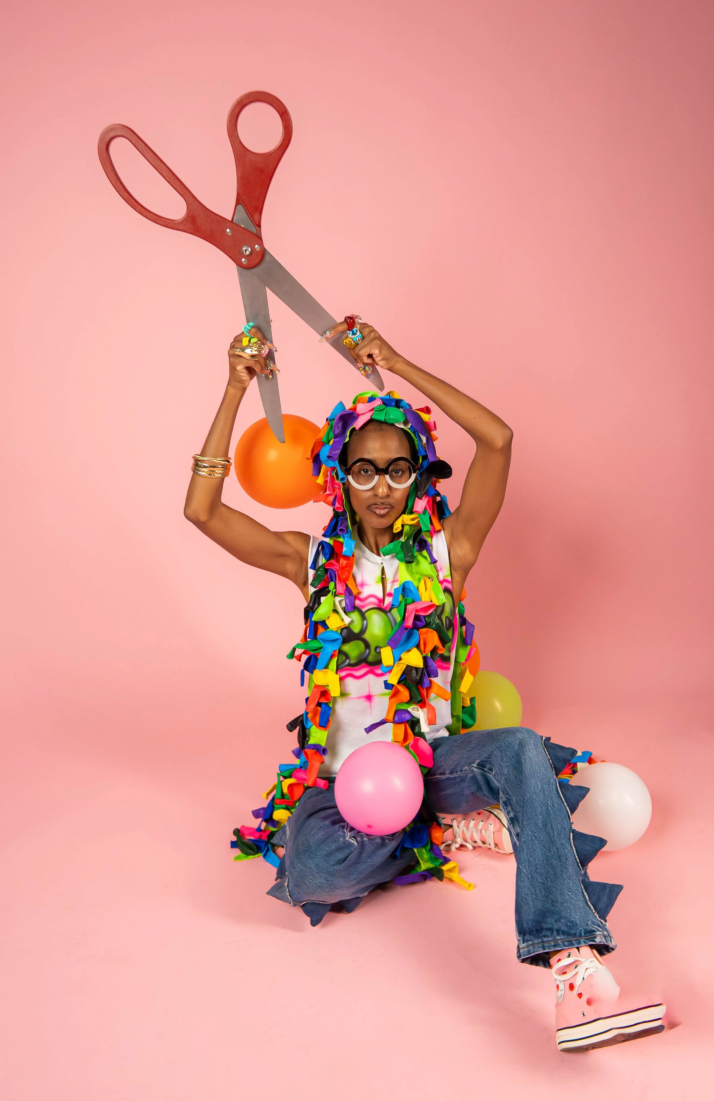 Person in colorful ribbon-covered clothing and glasses holding oversized scissors on a pink background with balloons