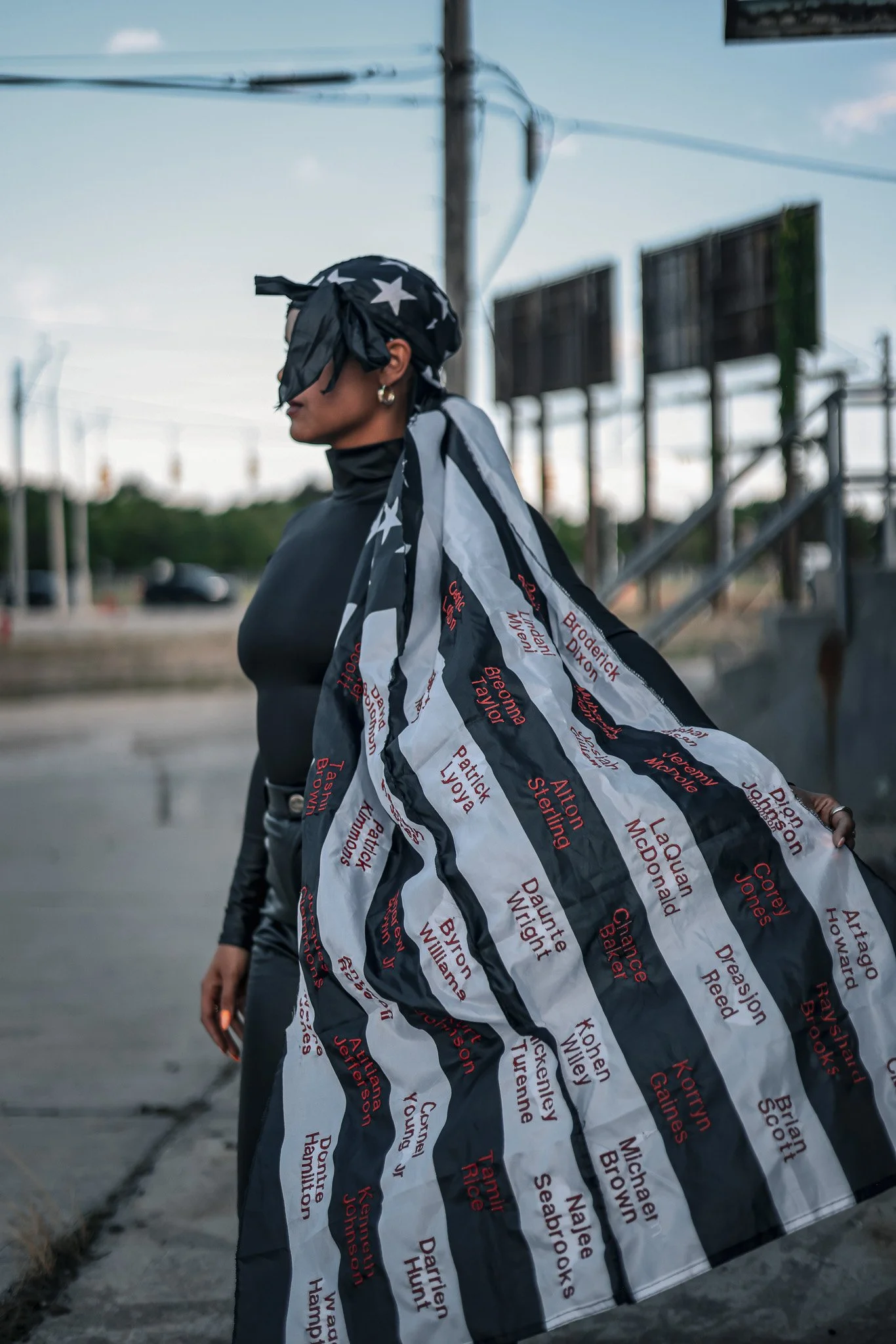 Person wearing a black-and-white striped cape printed with names and a matching star-patterned headscarf outdoors.