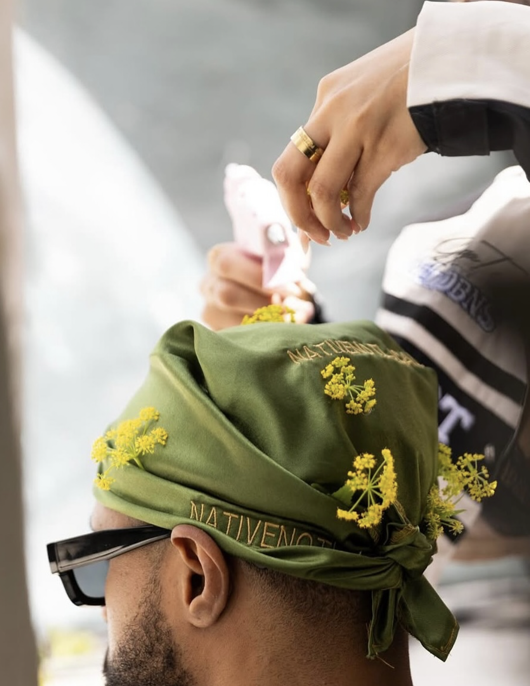 Close-up of a person wearing a green embroidered headscarf decorated with yellow flowers while another person adjusts it.