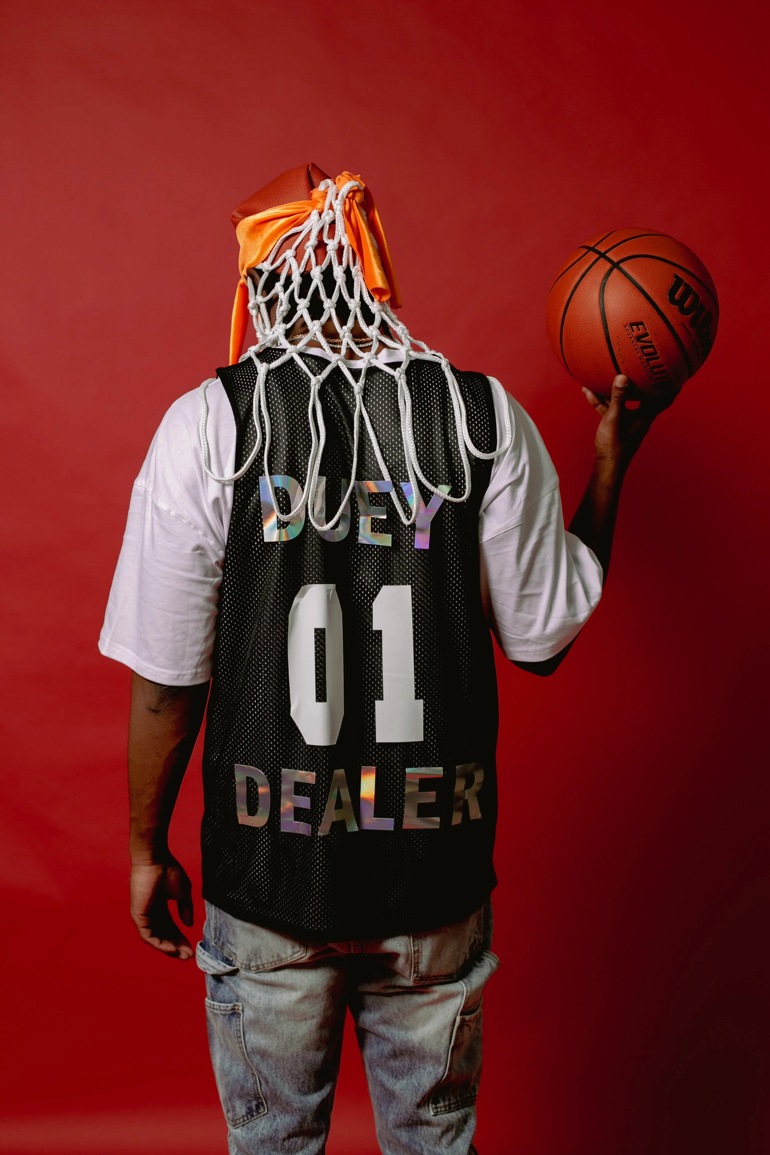 Basketball player wearing a hoop and net over an orange head covering, holding a basketball against a red background