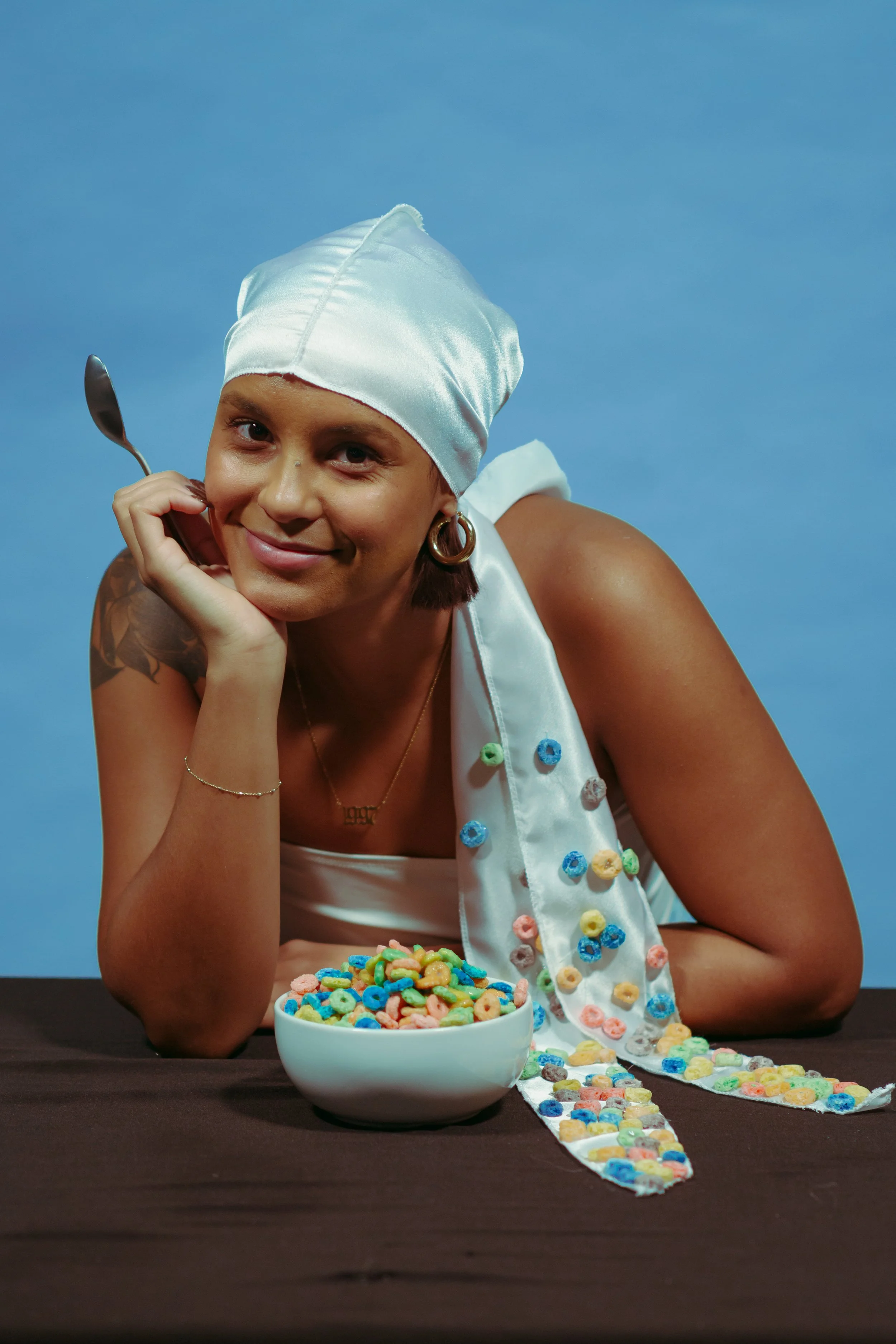 Smiling woman in a white headscarf holding a spoon beside a bowl of colorful cereal, with cereal scattered on her clothing and table