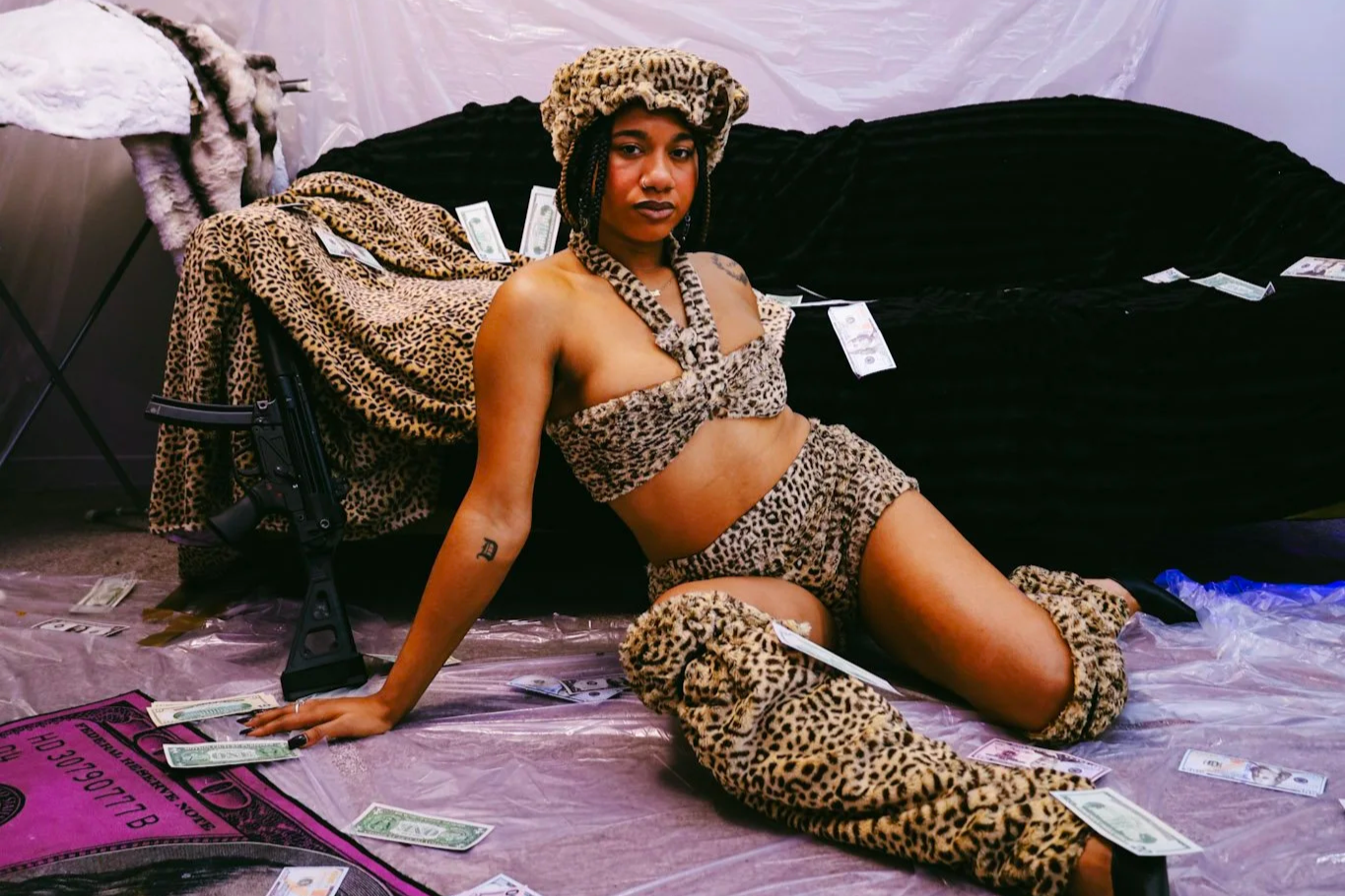 Woman in leopard-print outfit and furry hat posing beside a rifle, surrounded by scattered dollar bills.