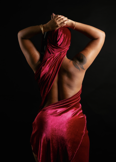 Back view of a woman wearing a red satin dress and headscarf, raising her clasped hands against a black background.