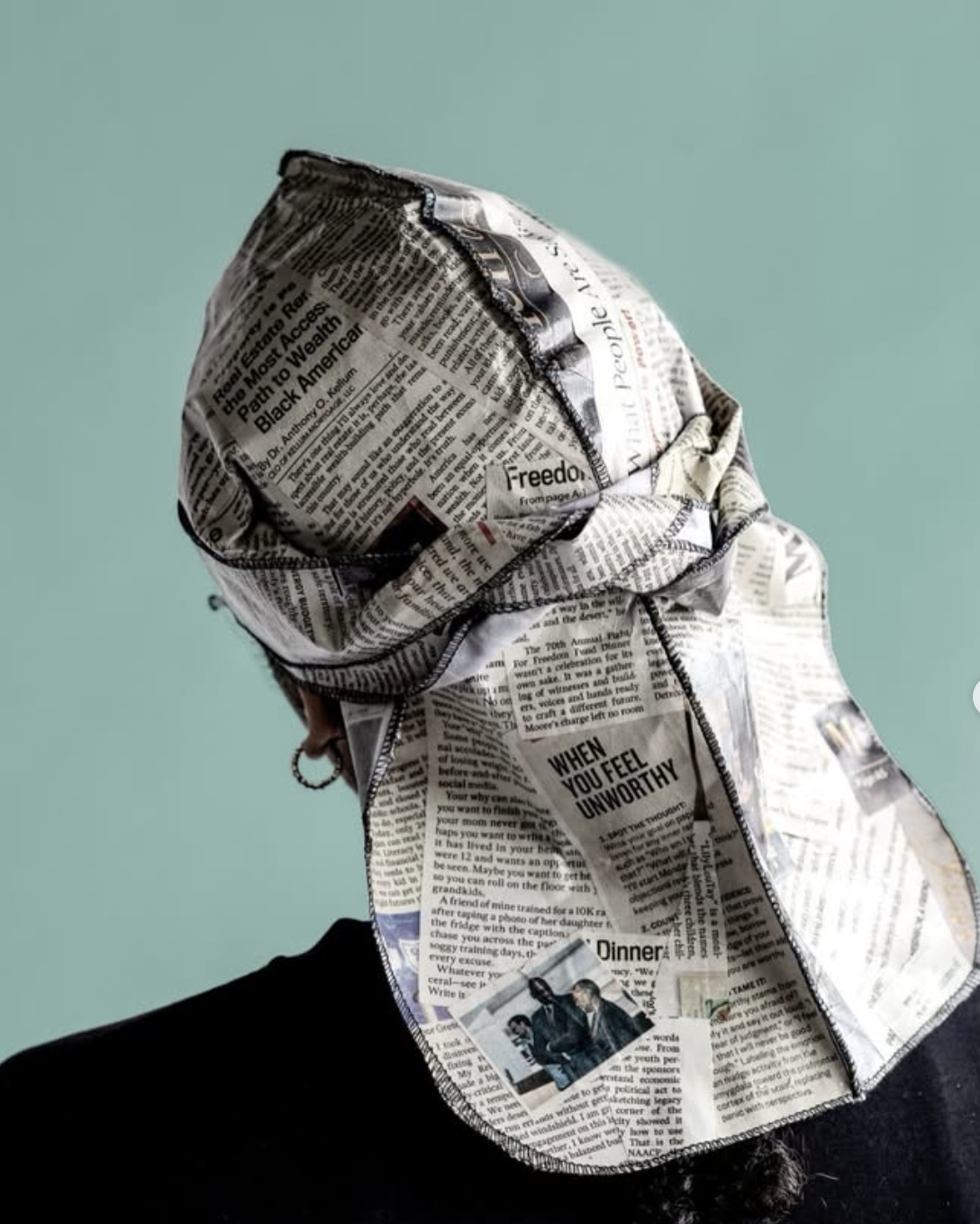 Person wearing a cap and hood made from stitched newspaper pages against a teal background