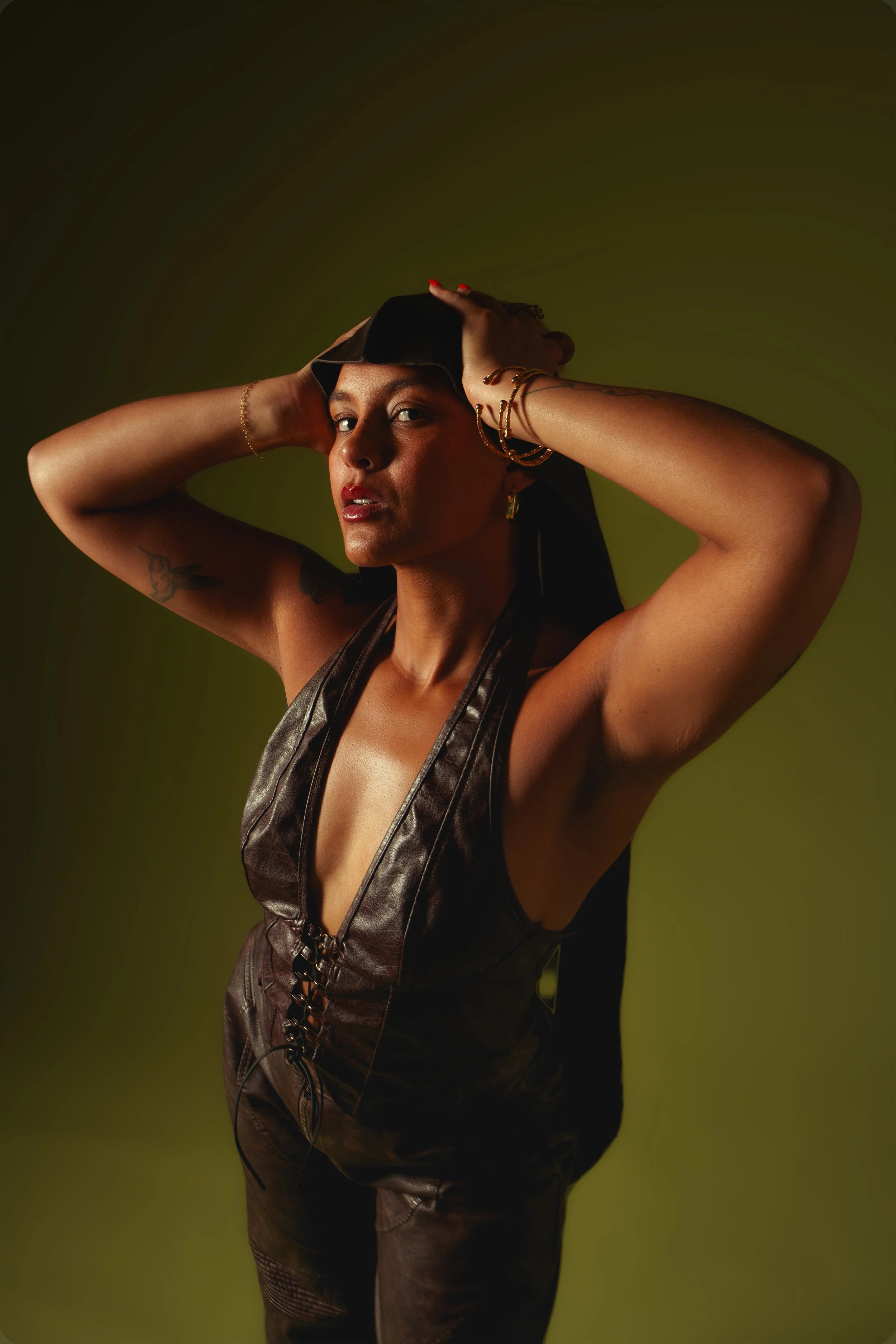 Portrait of a woman in a black leather outfit posing with both hands behind her head against an olive-green background