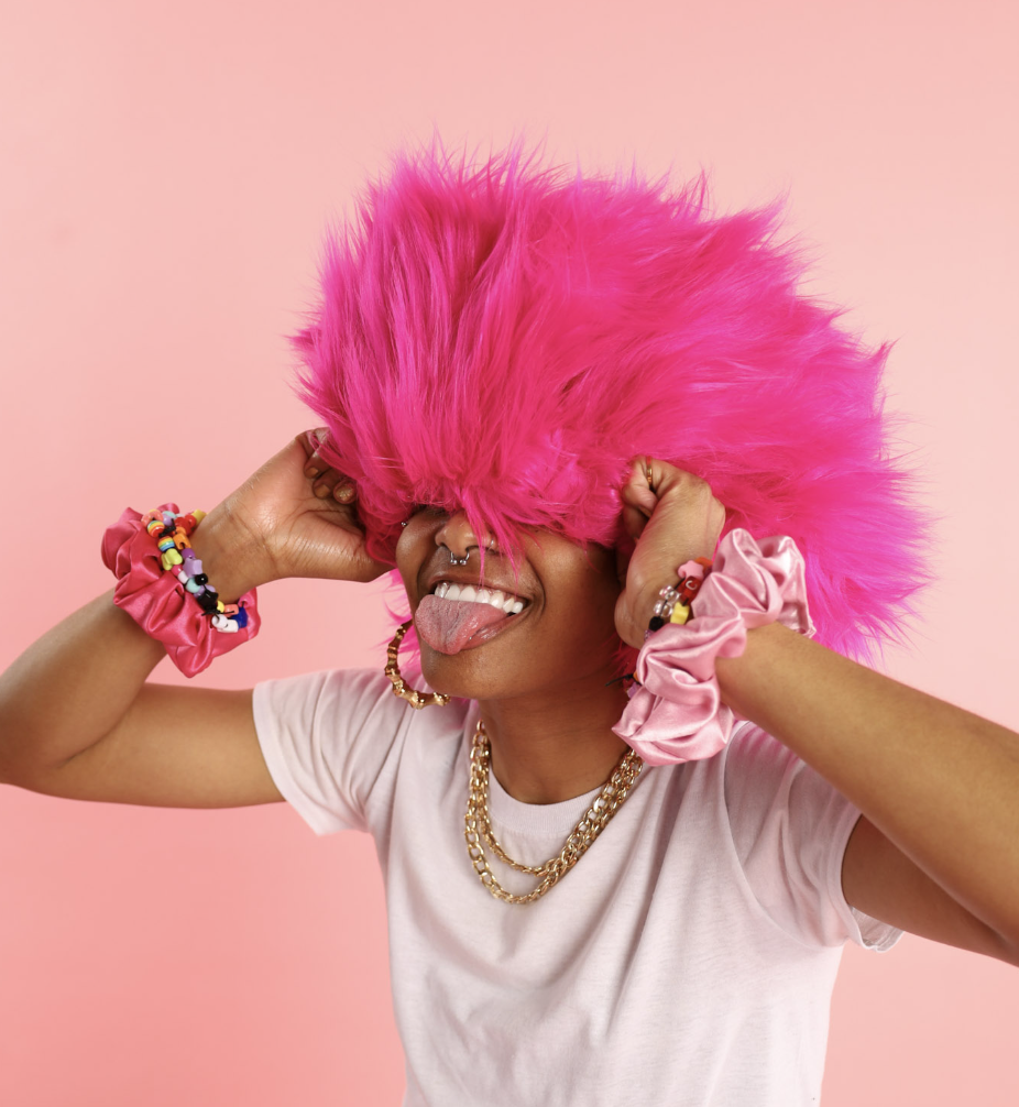 Person wearing a bright pink shaggy wig, sticking out their tongue, and holding the wig against a pink background