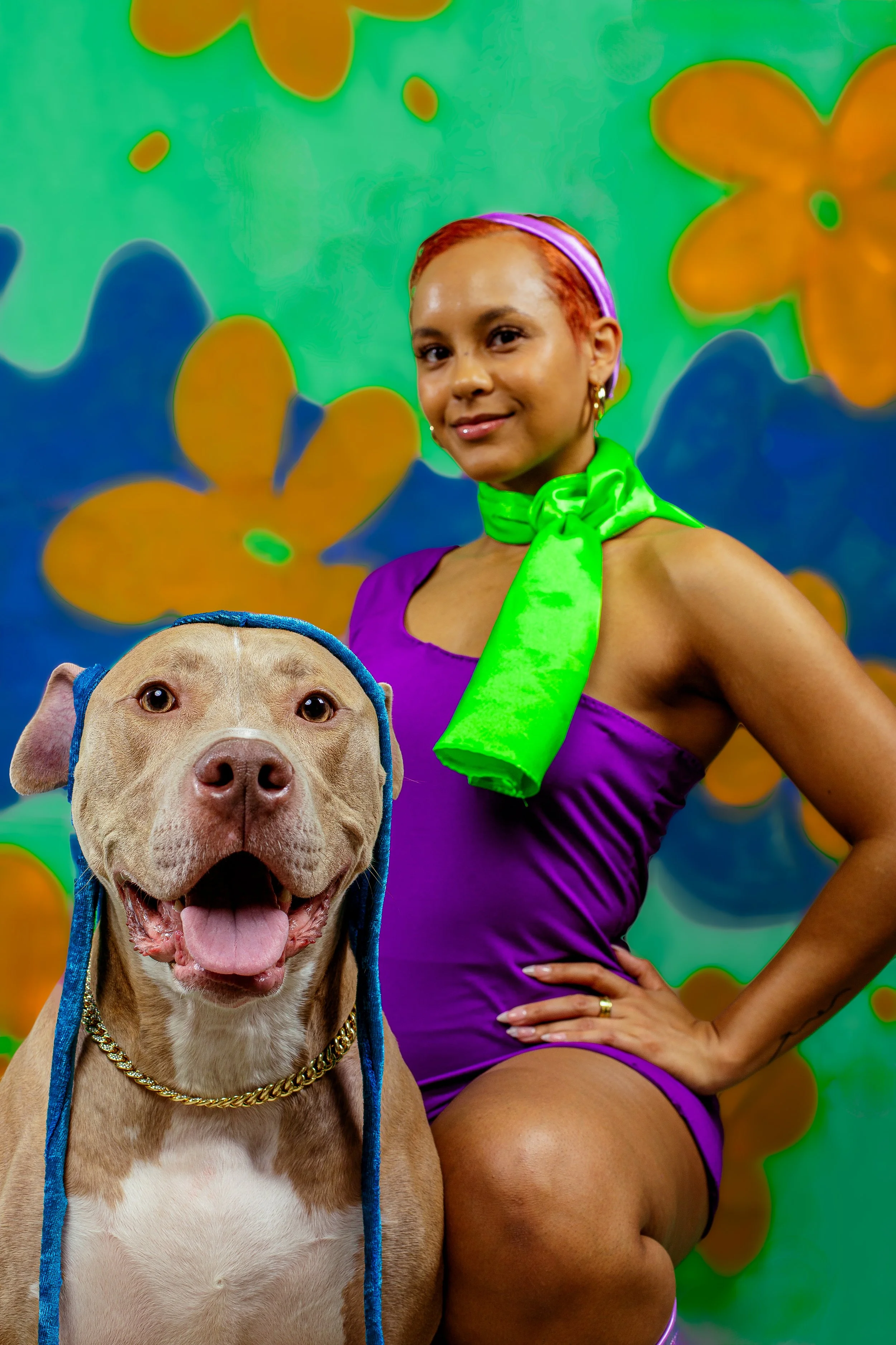 Woman in a purple outfit and neon green scarf posing with a smiling dog wearing a blue hood and gold chain against a colorful abstract background