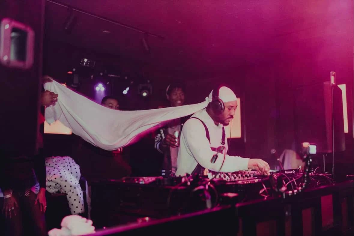 DJ performing on stage at a nightclub while people hold a stretched white sheet behind him