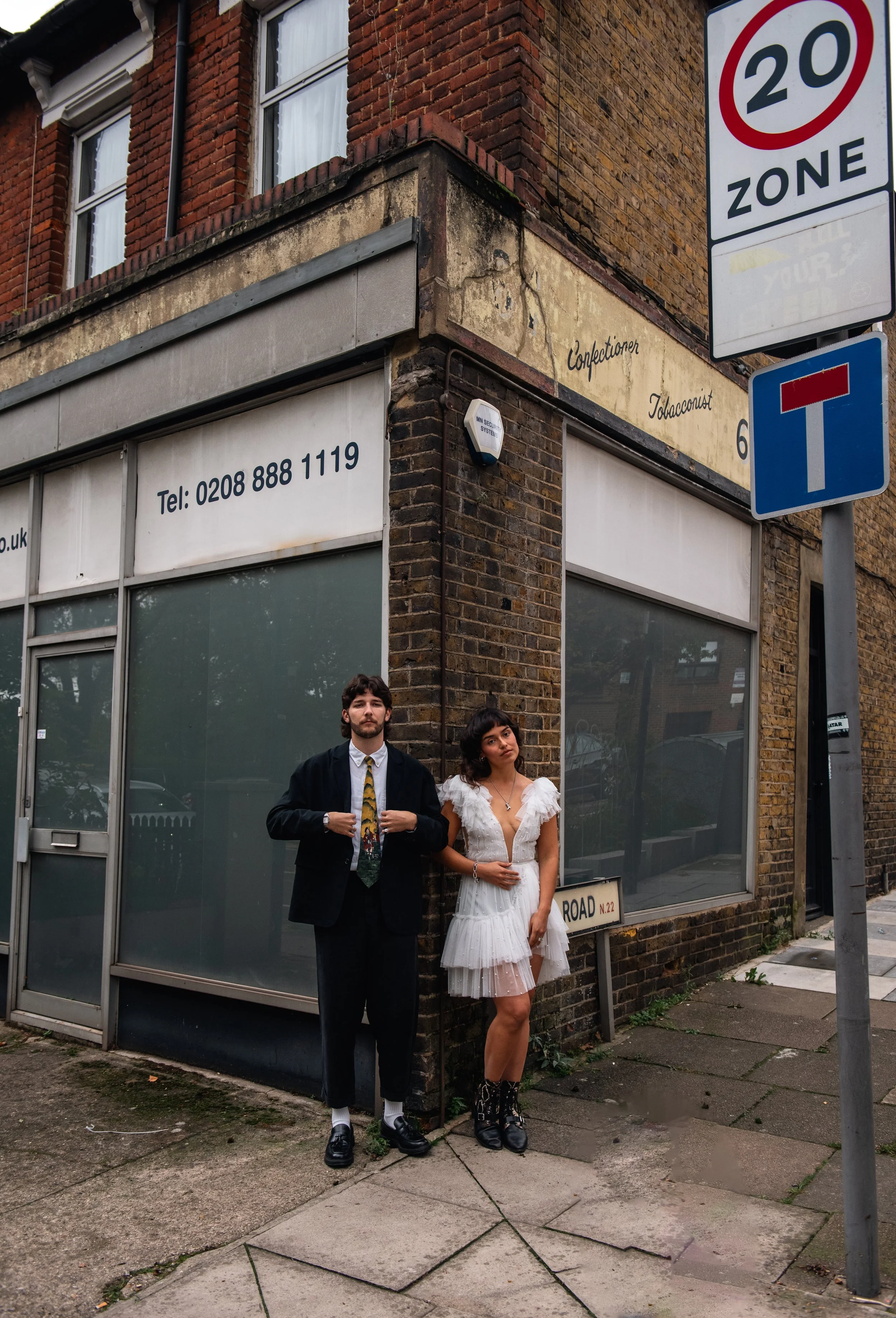 Two people standing on a sidewalk next to a brick building. The man is wearing a black suit and tie, and the woman is wearing a white dress and black boots. Street signs and building signs are visible.