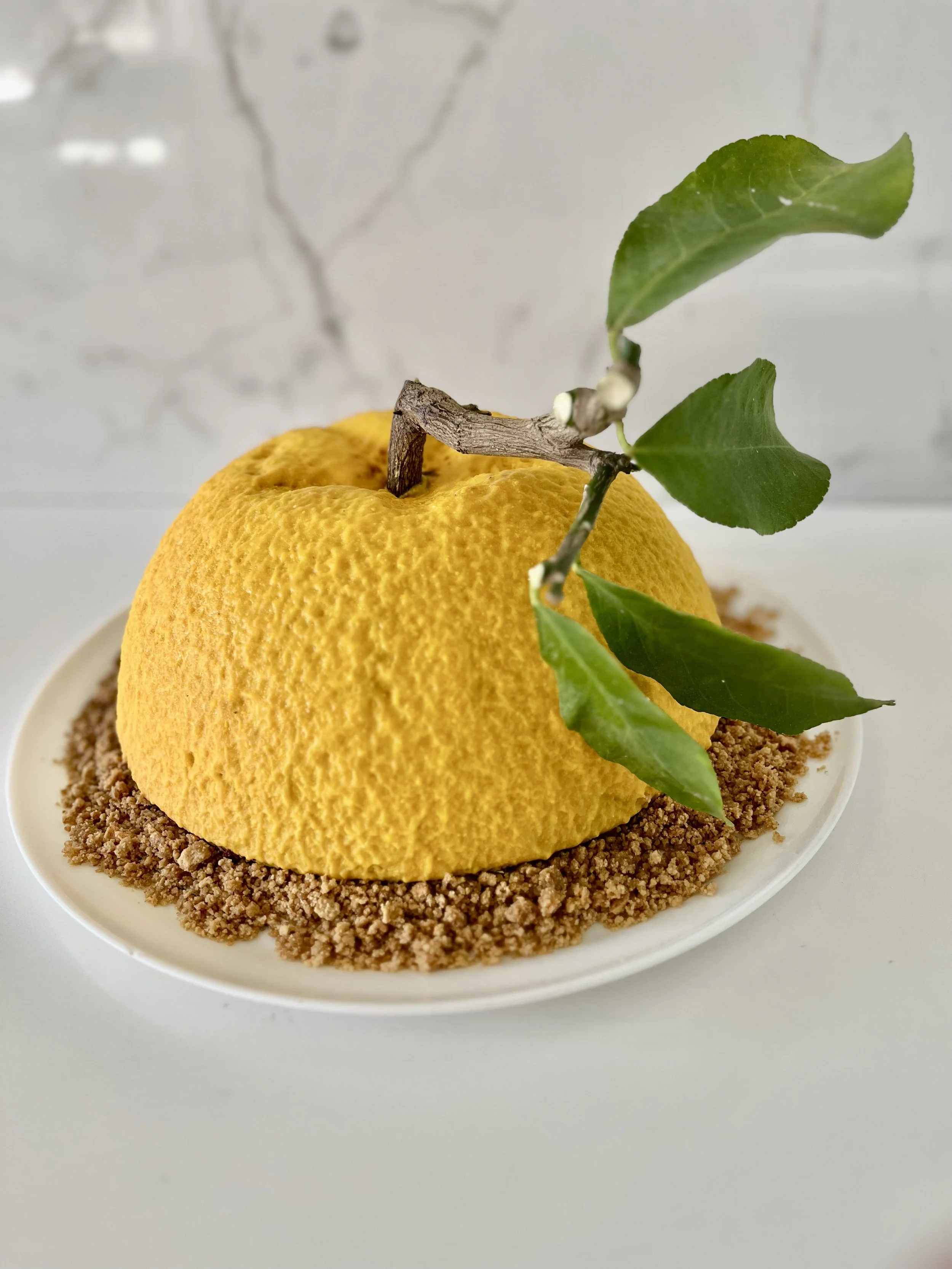 Nō 49 -------- YUZU MOUSSE CAKE - chocolate mousse interior with a cake and yuzu fruit jam, chocolate sponge, covered in a sprayed white chocolate textured crust peel.  Sitting on a lemon cookie crumble and lemon tree branch and leaves.
S,M, & large 