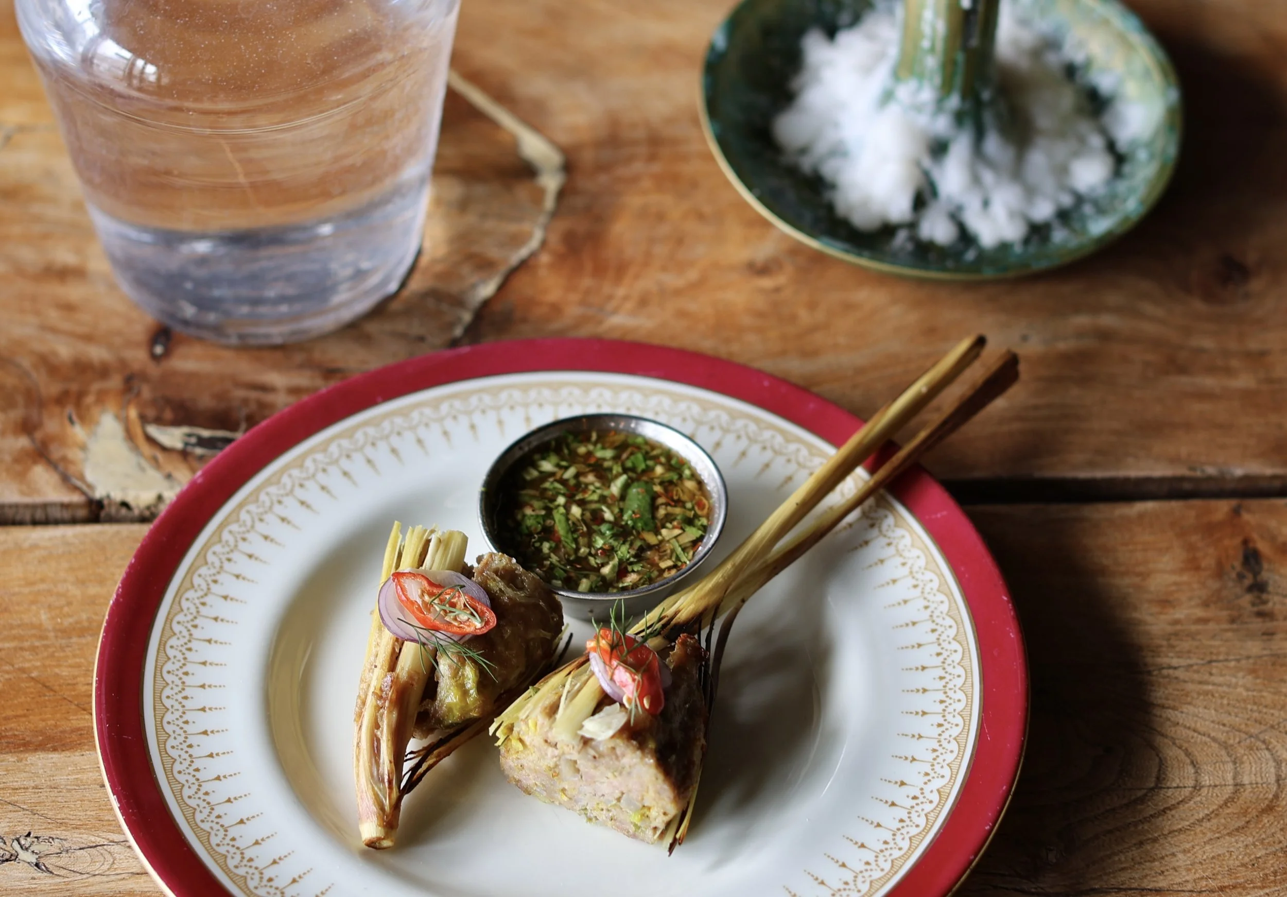 Vietnamese dish with rice paper rolls, herbs, and dipping sauce on a fancy plate, with a glass of water and a bowl of rice noodles in the background.