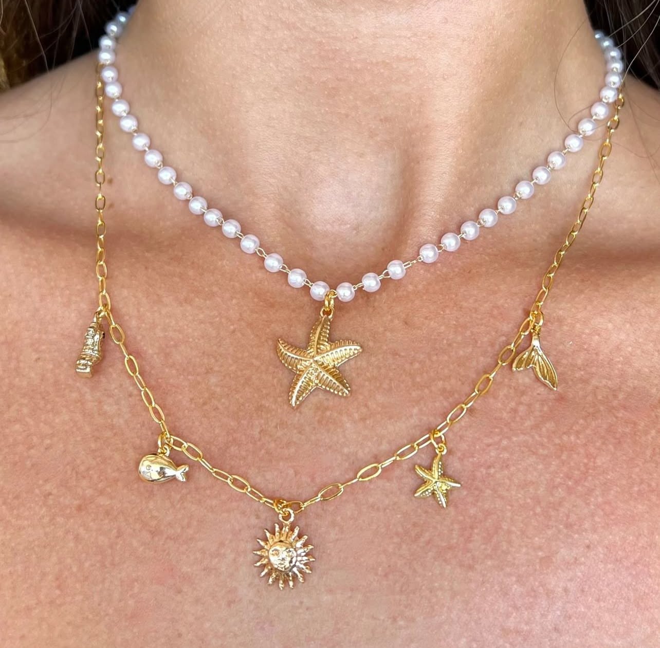 Two layered gold and pearl necklaces with starfish, fish, whale, and sun charms worn on a person's neck.