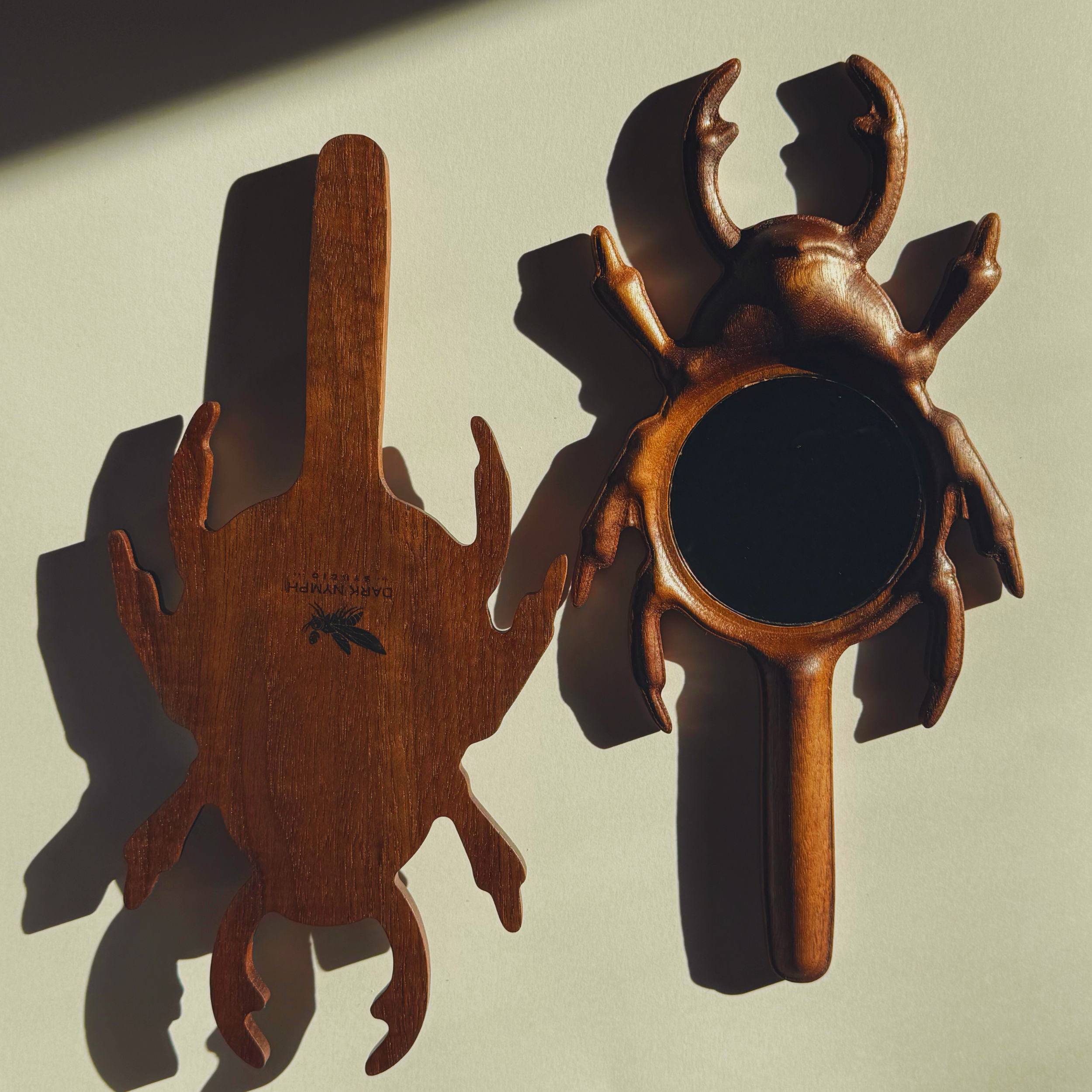 Two beetle shaped hand mirrors made out of wood with Dark Nymph Studio engraved on the back of the first one