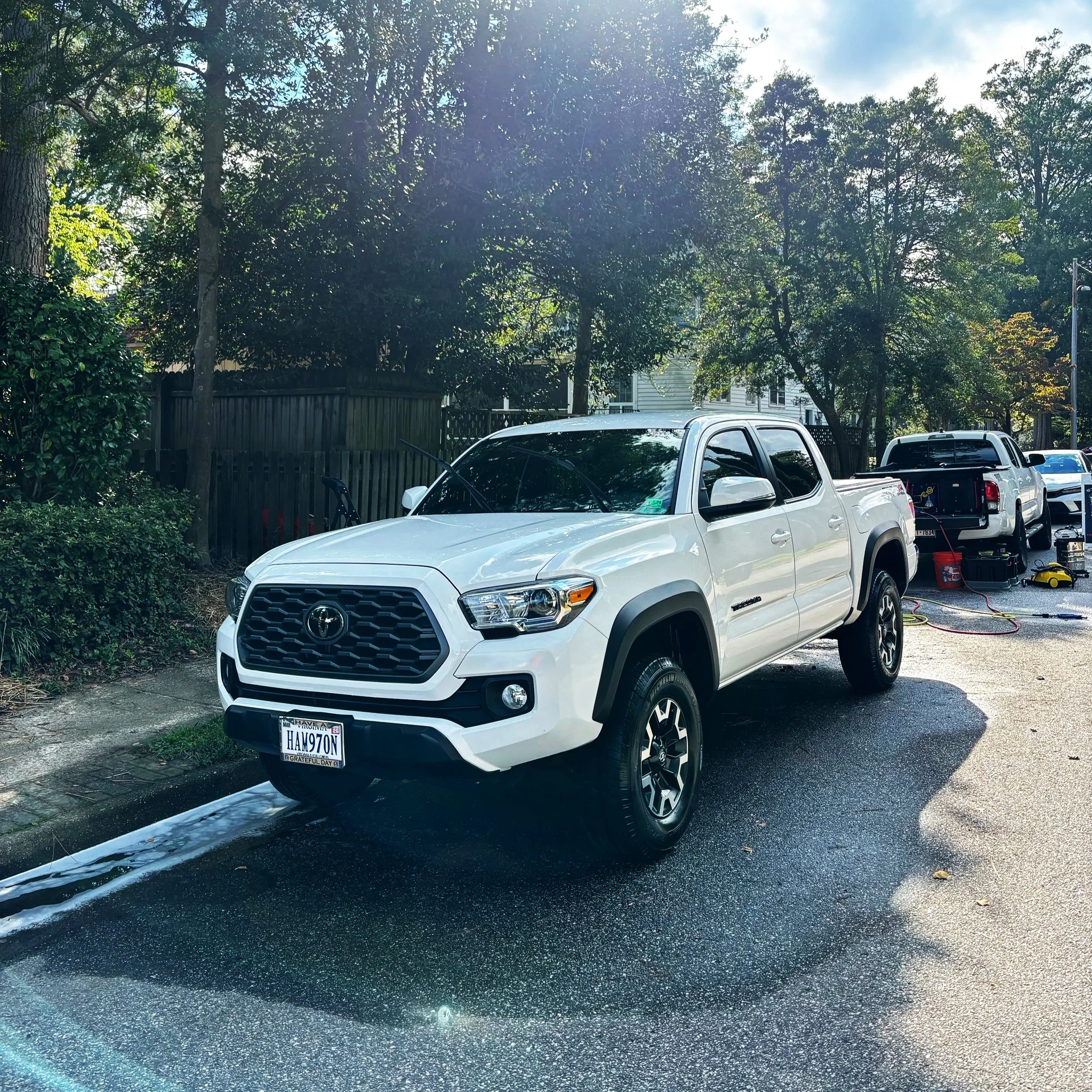 White Toyota Tacoma parked on a wet street with trees and houses in the background, and car repair equipment nearby.