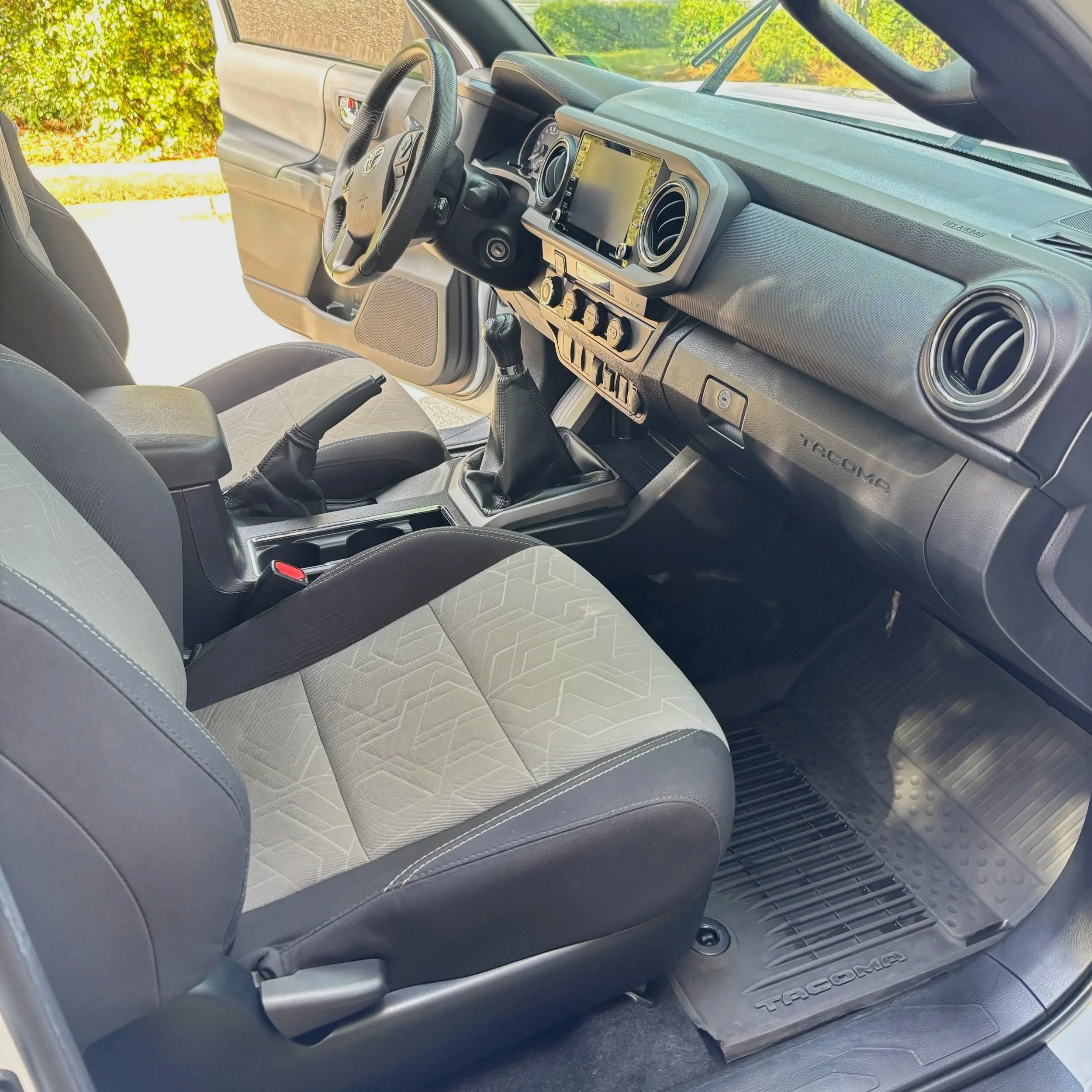 Interior of a Toyota Tacoma truck, showing the driver's seat, steering wheel, dashboard with a touchscreen display, manual gear shift, and front passenger seat, with sunlight outside visible through the open door.