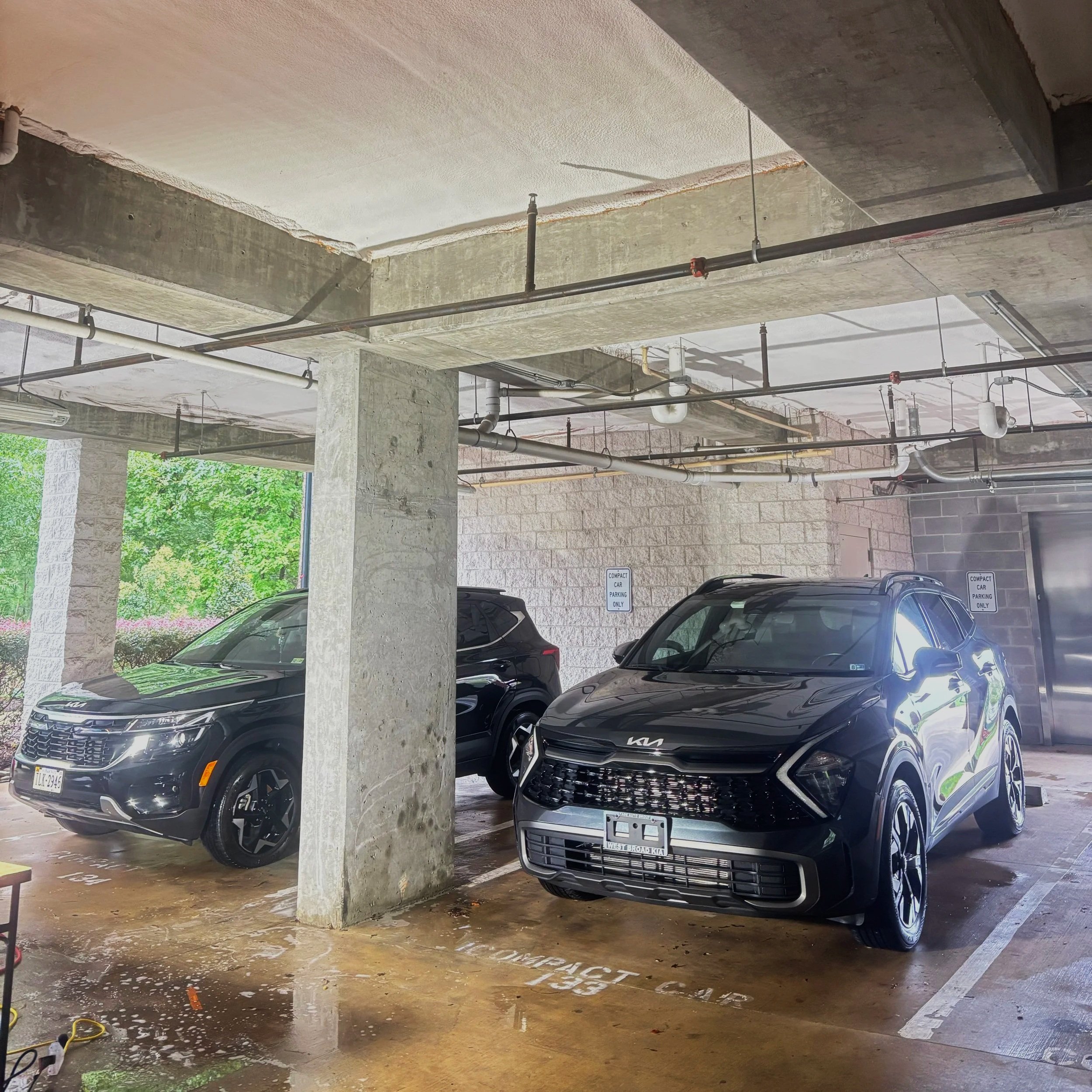Indoor parking garage with two black Kia vehicles, concrete pillars, exposed ceiling pipes, and a view of green trees through the open side.