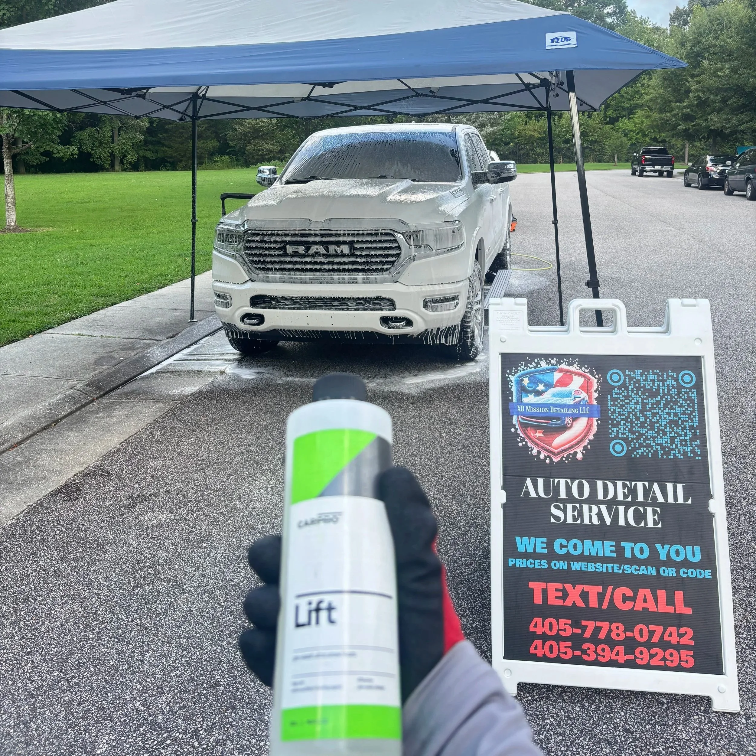 A white Ram truck being washed under a blue canopy at an auto detail service station. Someone is holding a spray bottle labeled 'Lift' in the foreground, and a sign with contact information is visible.
