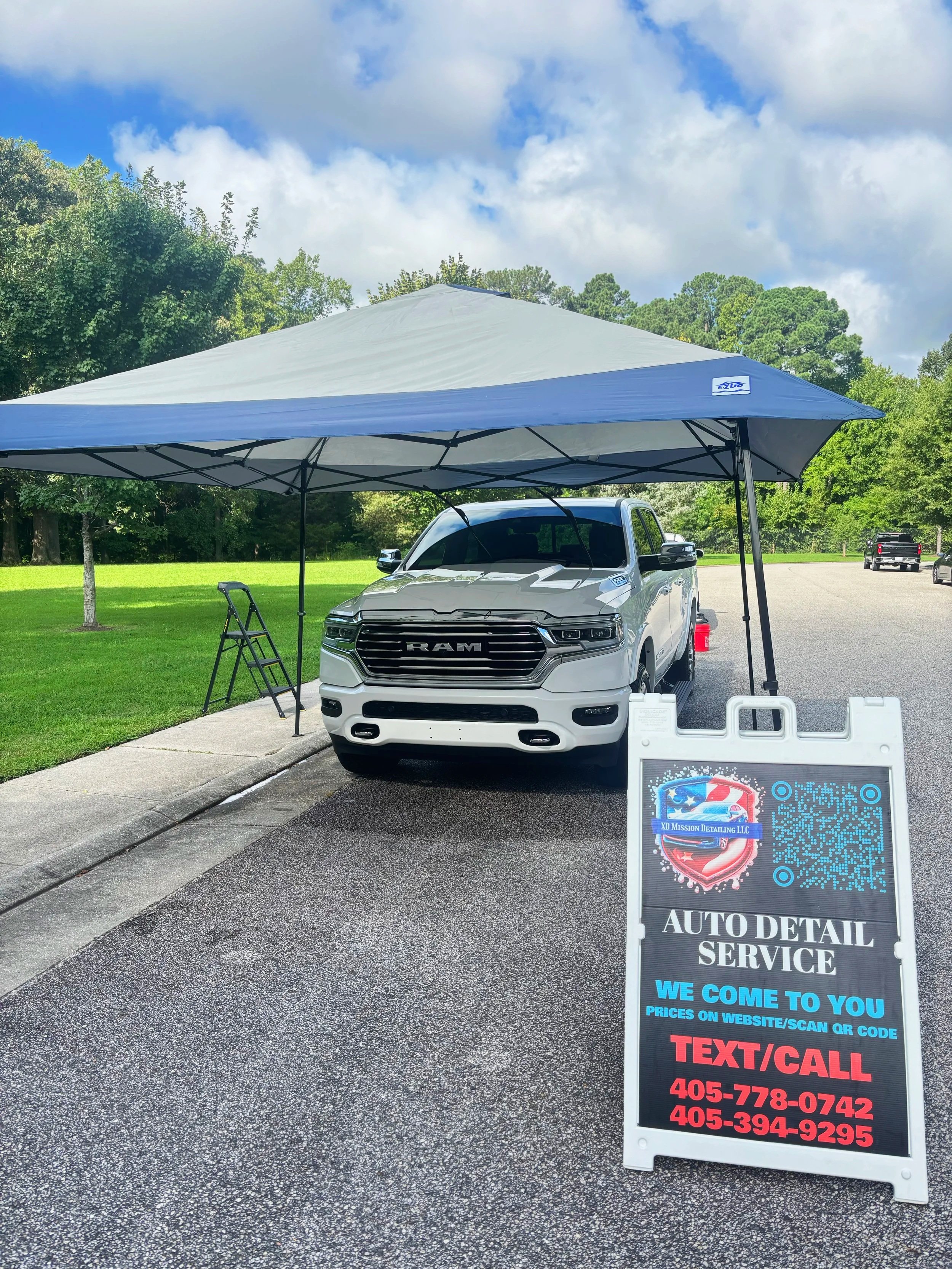 Auto detailing service booth with a white RAM truck parked under a canopy, a sidewalk, trees, and a grassy area in the background. There is a signboard with contact information and QR code for the service.