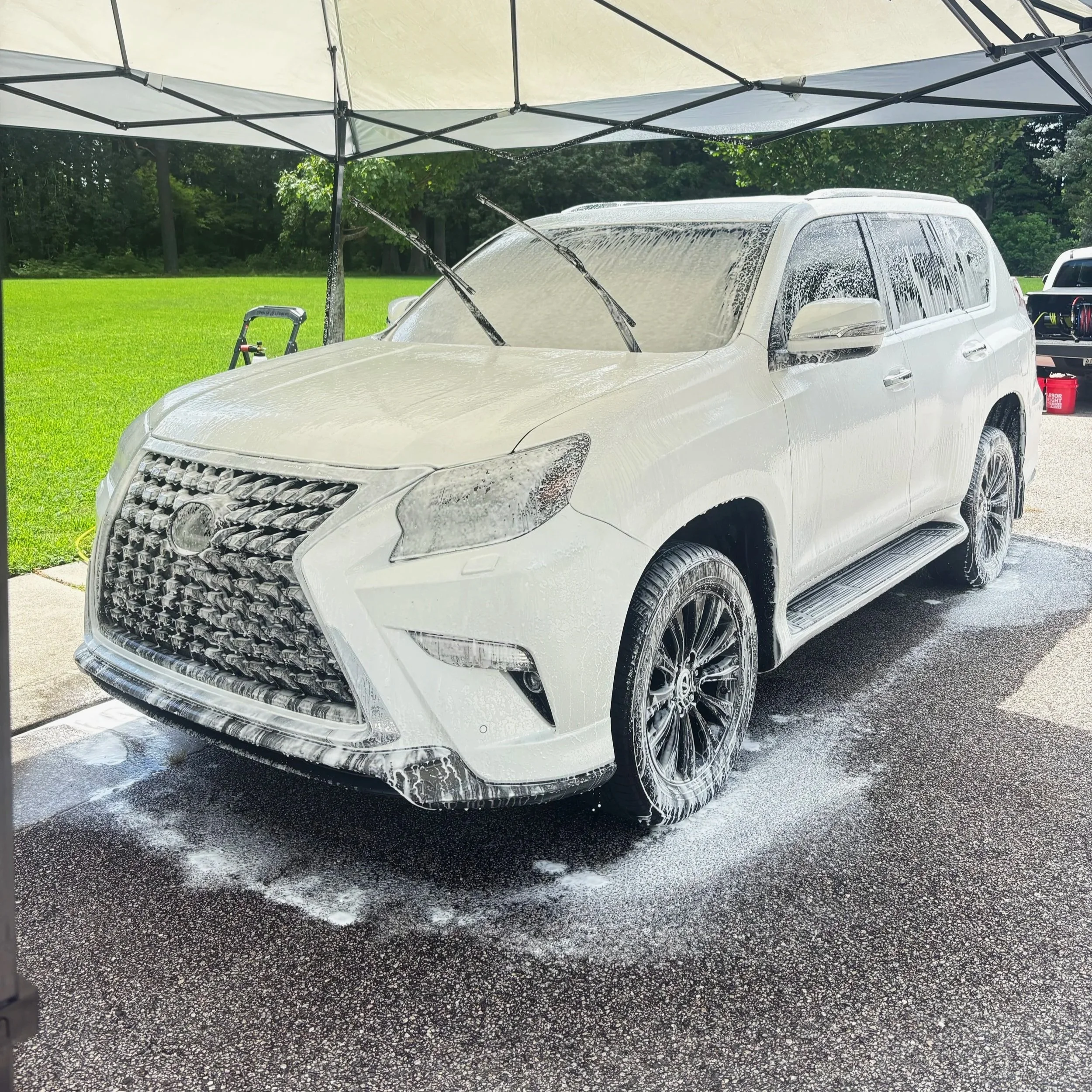 White Lexus SUV covered in soap and foam during a car wash, parked on a driveway under a canopy with a grassy lawn and trees in the background.