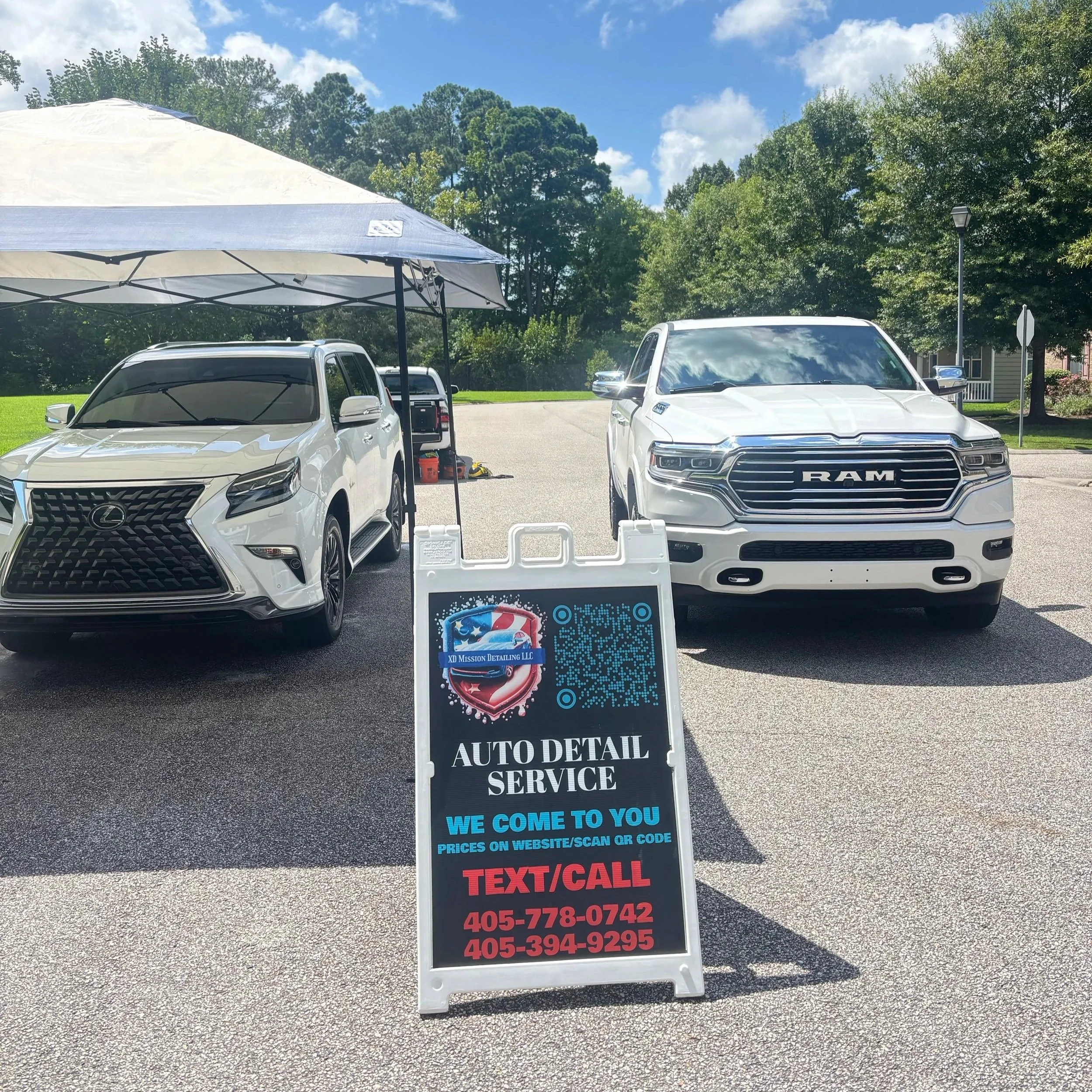 A white Lexus SUV and a white RAM truck parked outdoors under a large canopy tent on a sunny day with trees in the background. A signboard in front advertises auto detailing services, with a QR code, contact phone numbers, and the message 'WE COME TO