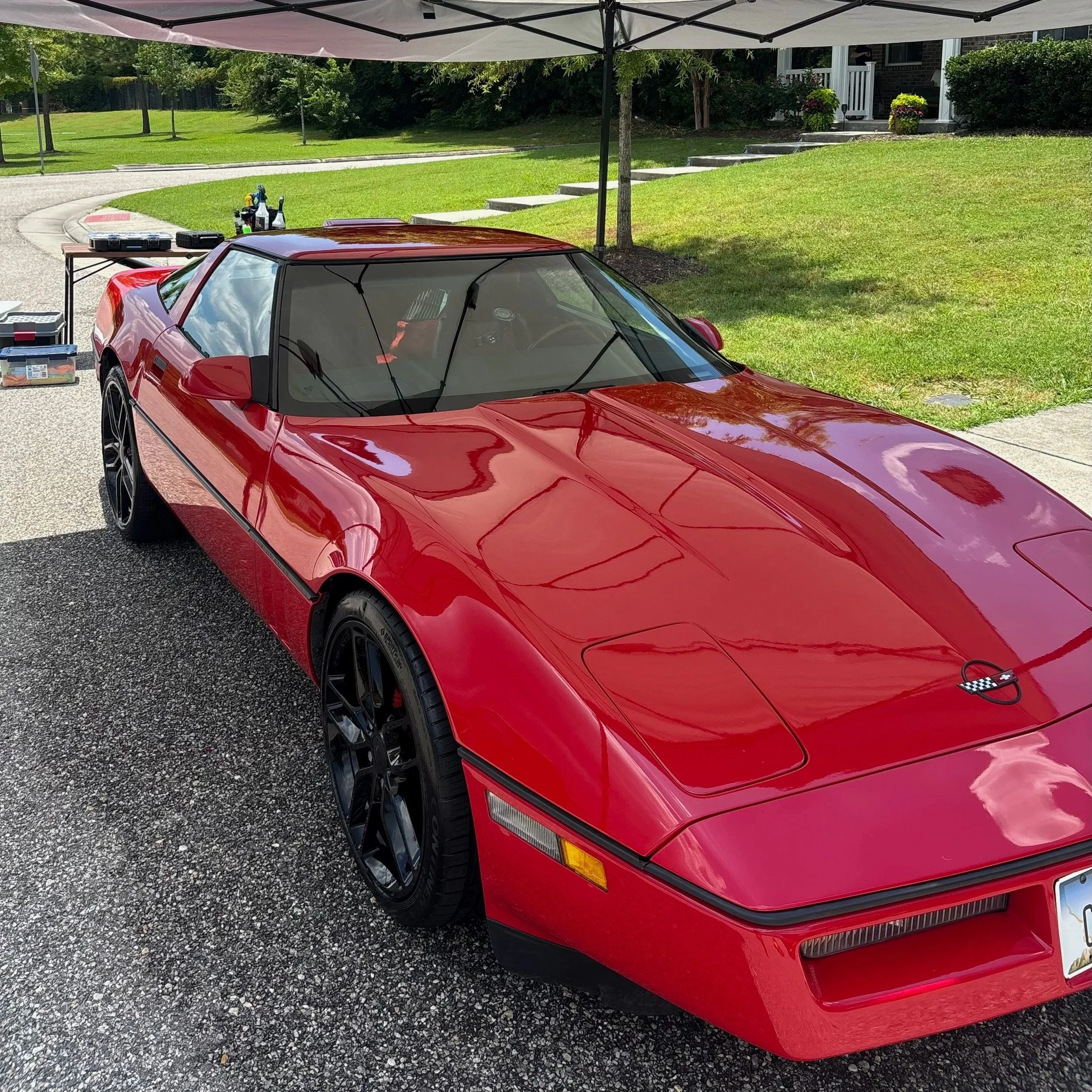 Red Corvette sports car parked under a canopy in a driveway with tools and equipment nearby, a grassy lawn and houses in the background.