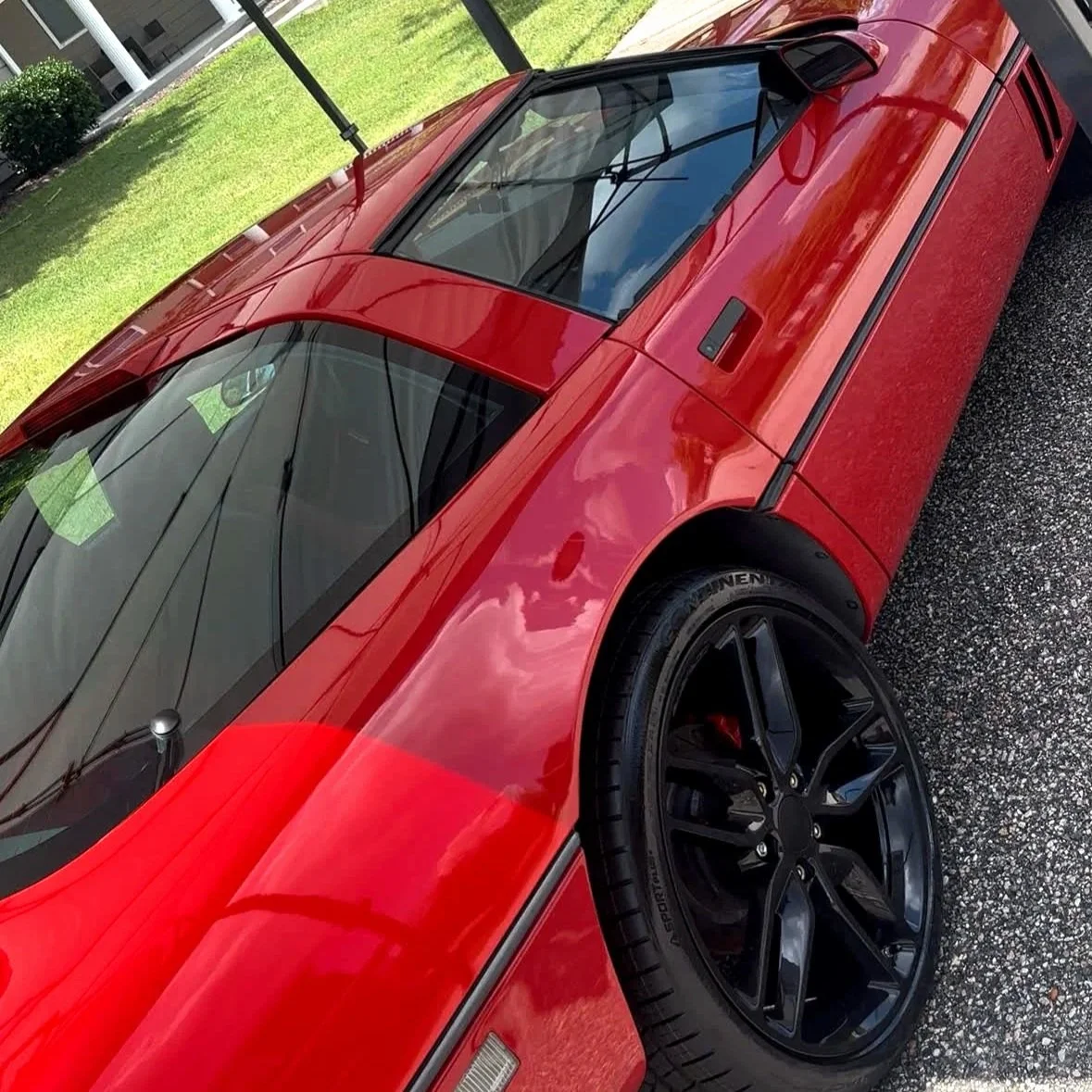 Red sports car with black wheels parked on a gravel surface near a grassy area.